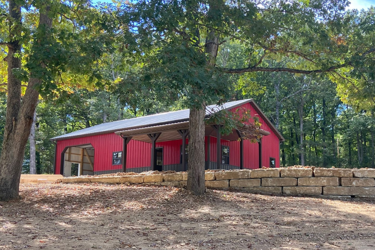 Red barn with covered entrance, set against a backdrop of trees, with a low stone wall in the foreground.