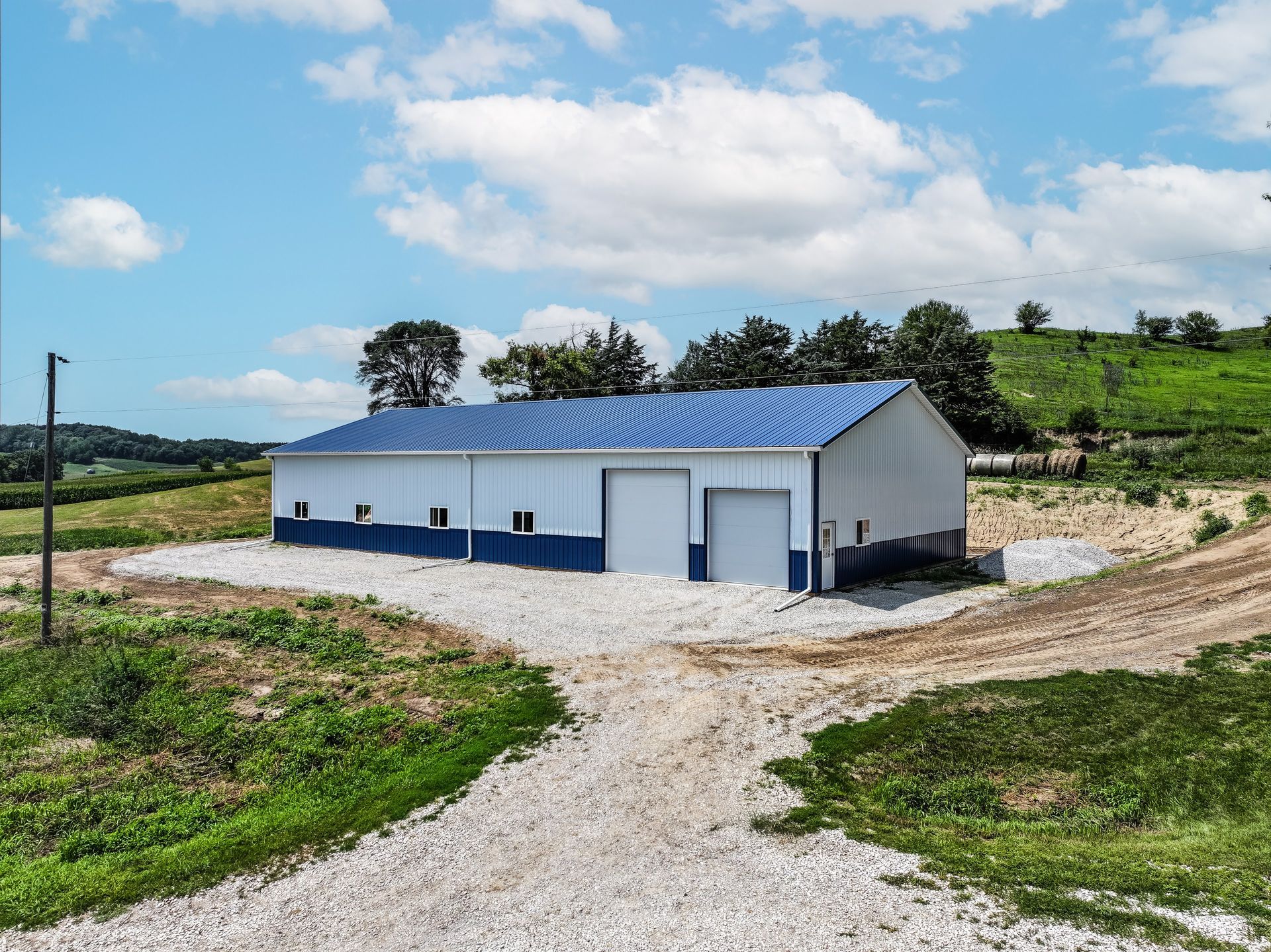 Blue and white metal building on a gravel lot, rural setting with blue sky and green hills.