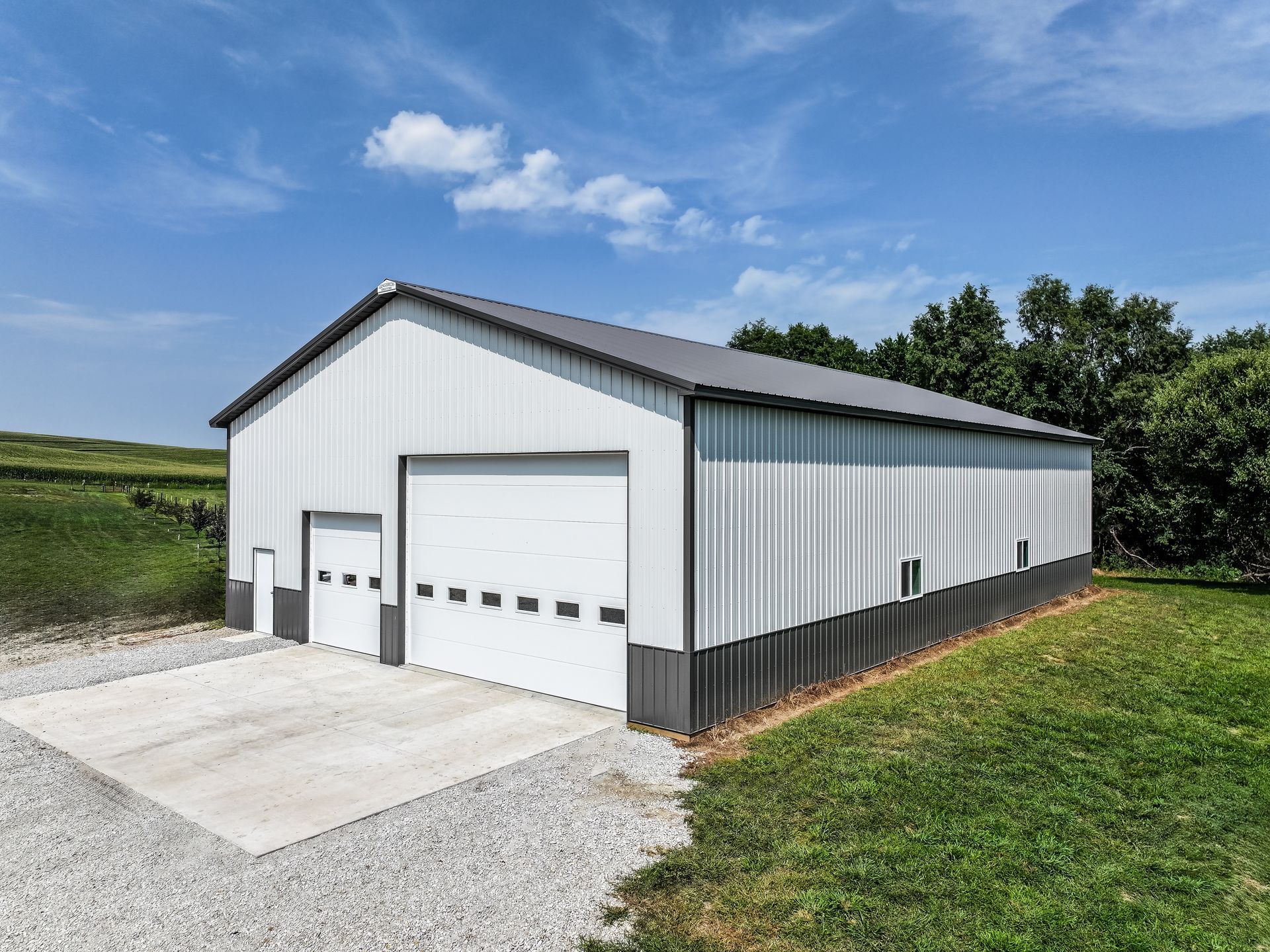 White and gray metal barn with large garage doors, gravel driveway, and green field.