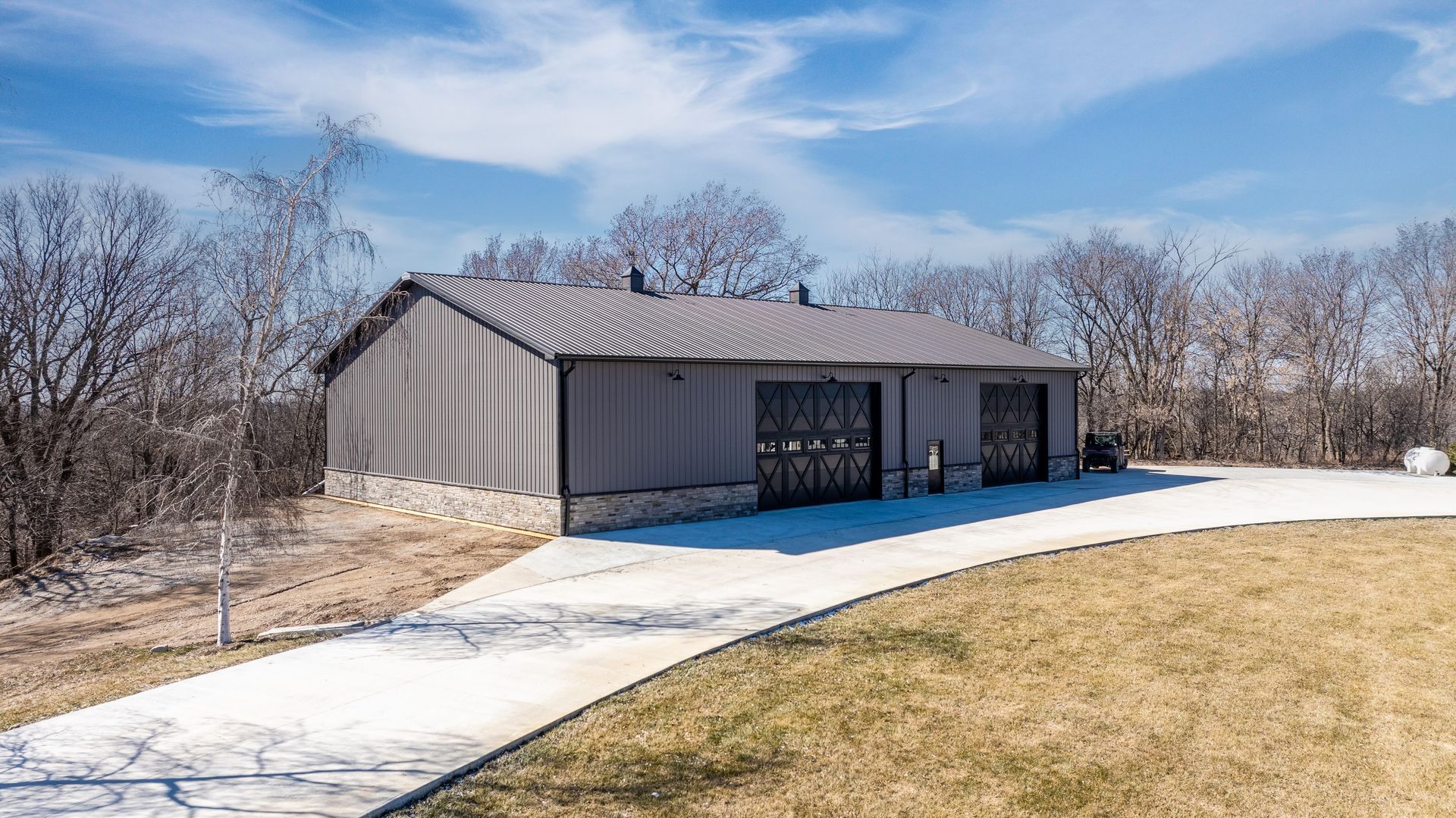 Gray garage with two bays, set on a concrete driveway, against a backdrop of trees and a blue sky.