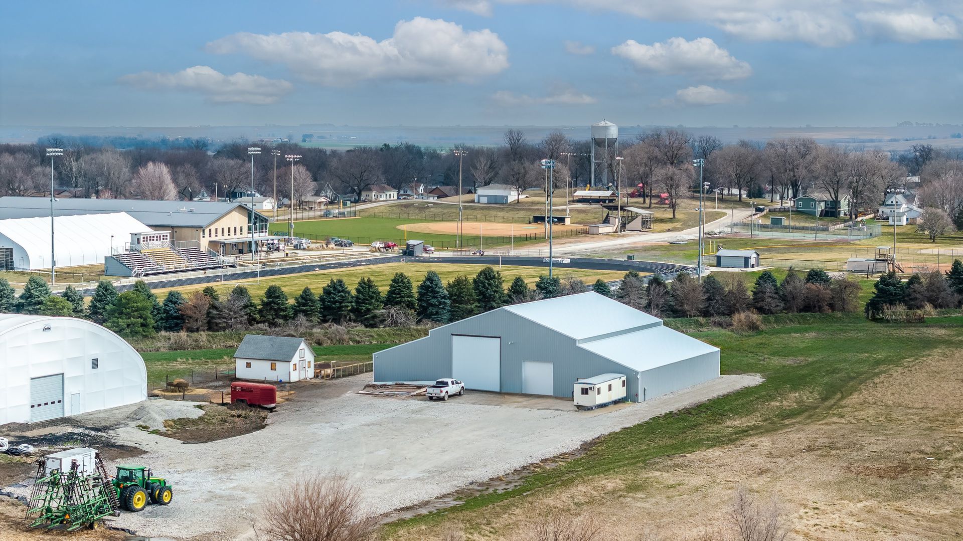 Aerial view of a farm with large storage buildings, a tractor, and a town in the background under a cloudy sky.