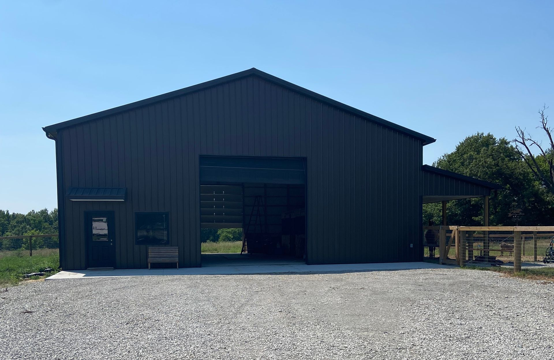 Dark green metal barn with open bay door, gravel driveway, and small side entrance.