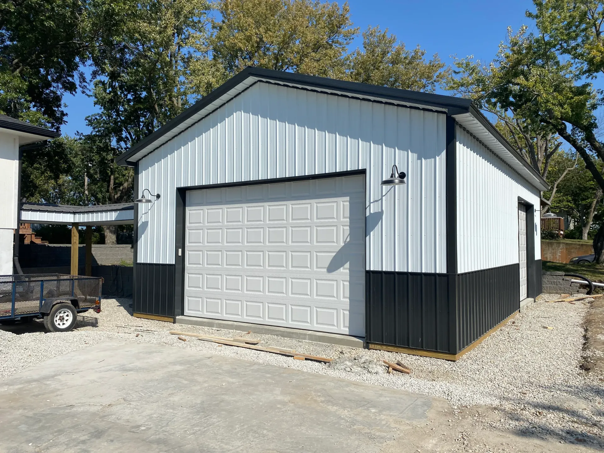 White and black metal garage with closed gray garage door; gravel driveway.