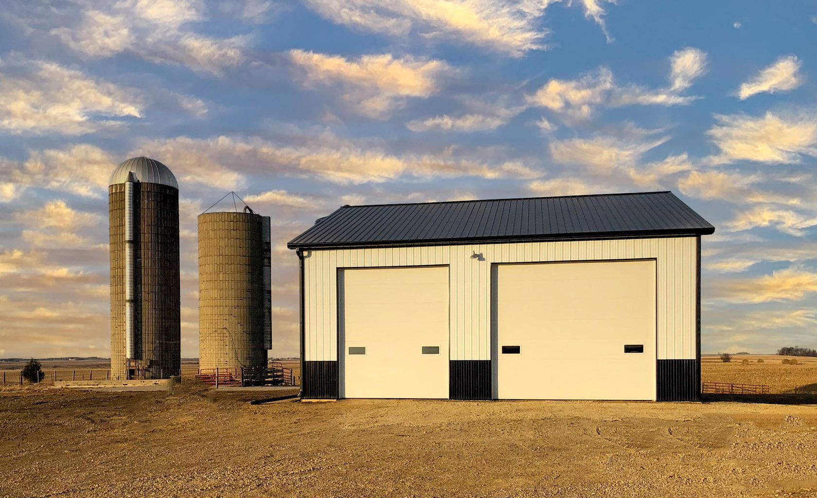Farm with two silos and a white garage under a colorful sky.