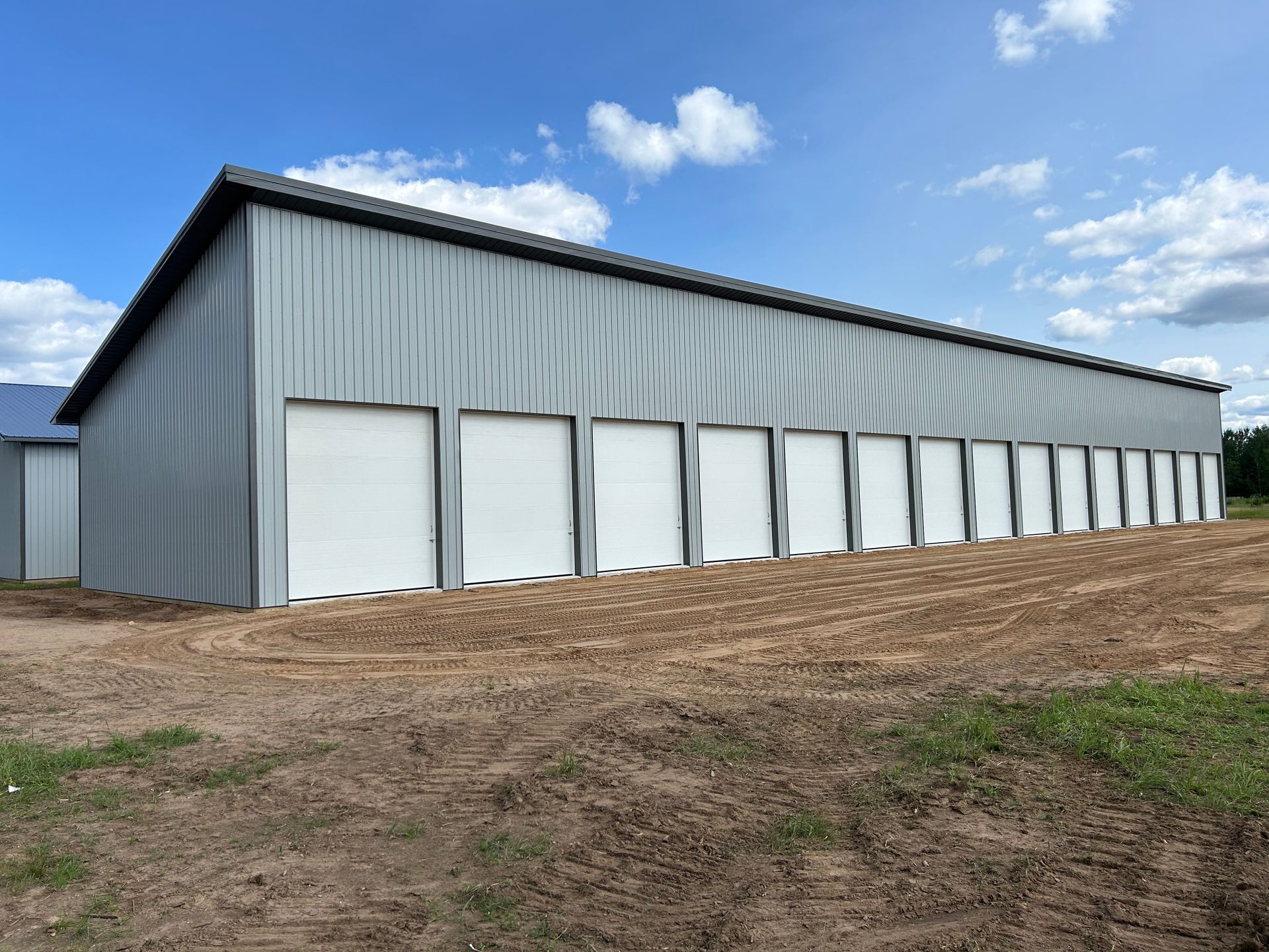 Metal-sided building with brown roof and trim, a porch, and a large garage door. Green grass, trees, and a fence.