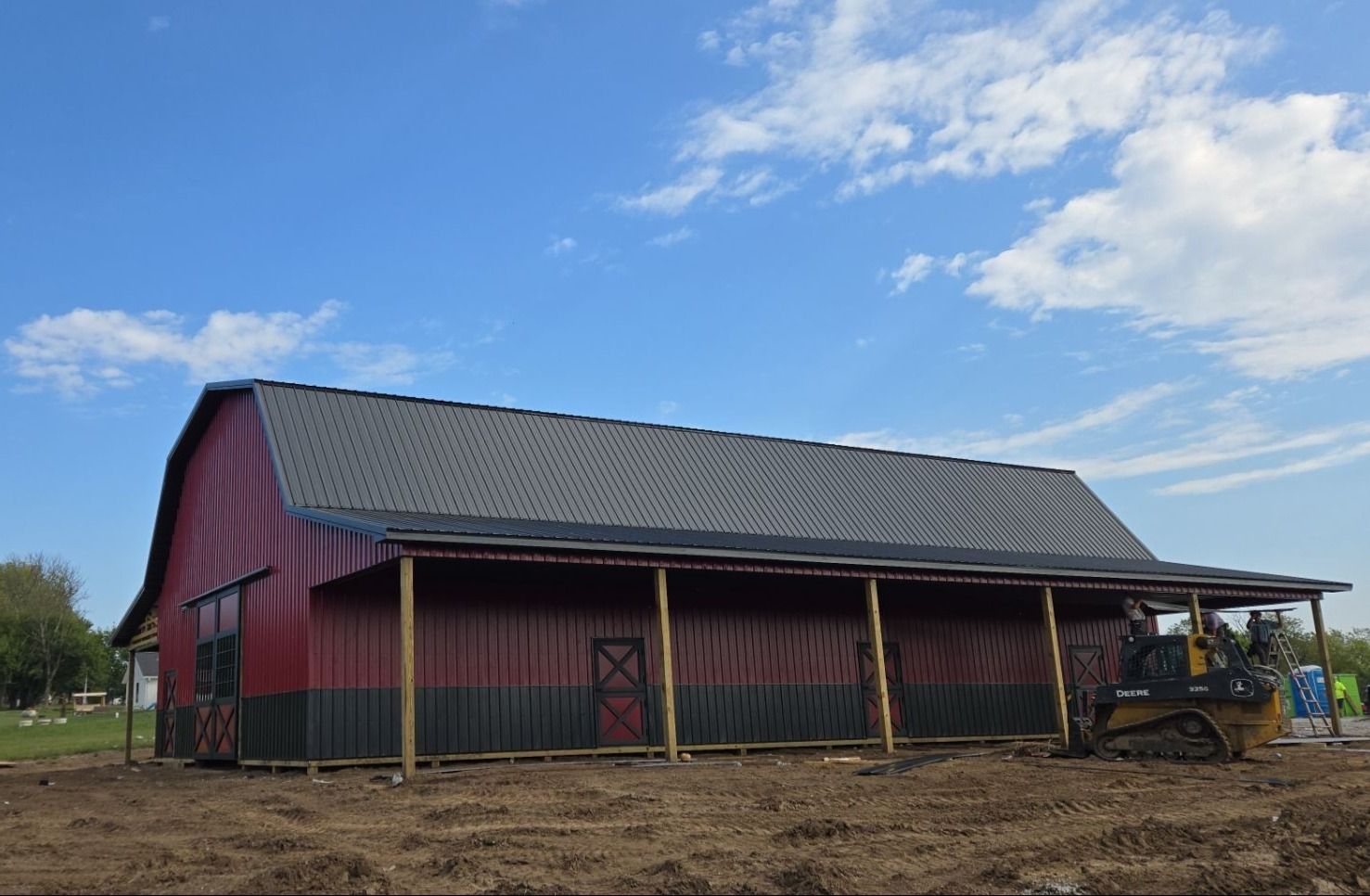 Red and black barn with a gray roof and a covered porch. A bulldozer is parked nearby under a blue sky.