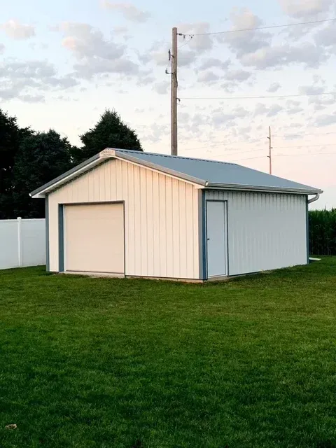 White metal garage with a gray roof, door, and garage door, set in a grassy yard with a utility pole in the background.