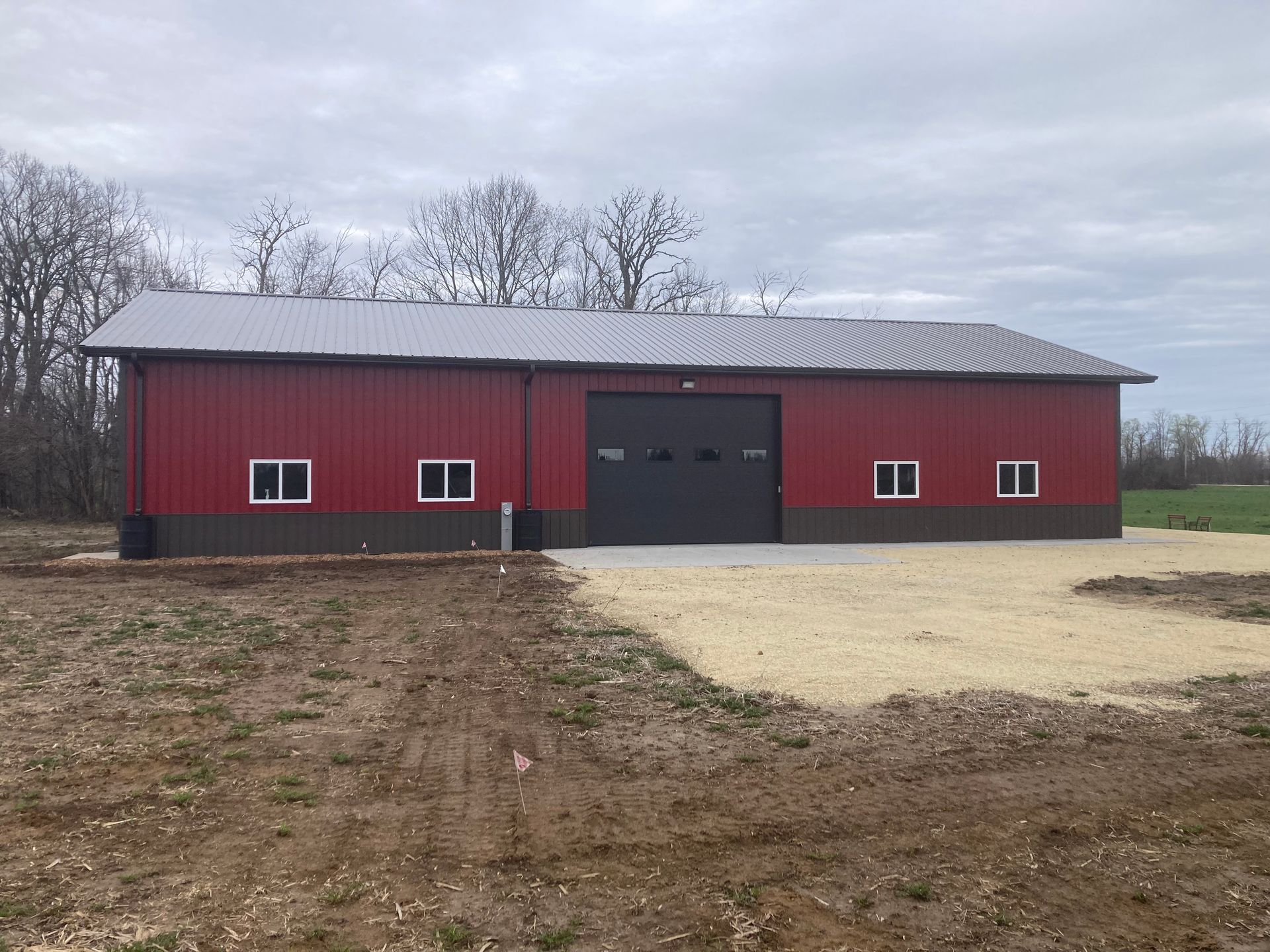 Red and brown metal barn with a large gray garage door and gravel driveway, under a cloudy sky.