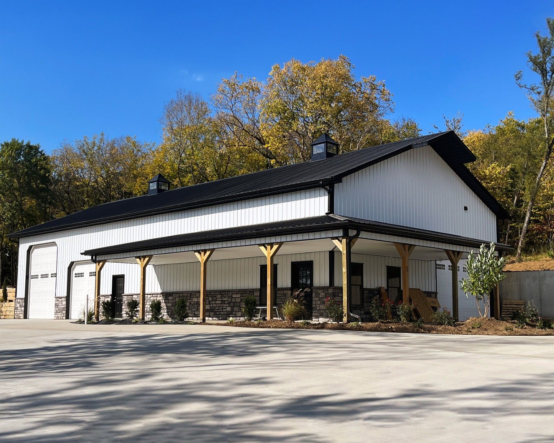 White barn with black roof and stone accents, blue sky, fall foliage.