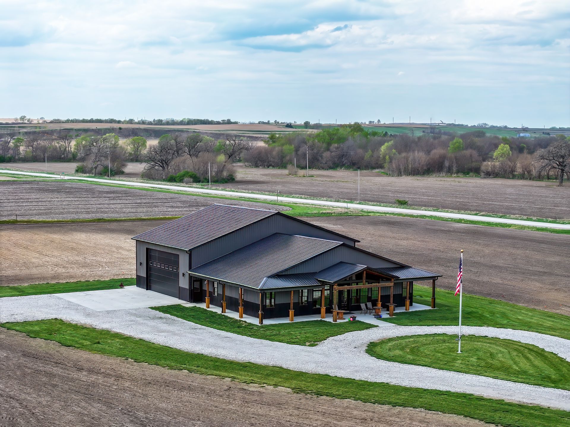A dark gray modern home with a covered porch, gravel driveway, and American flag in a rural field.