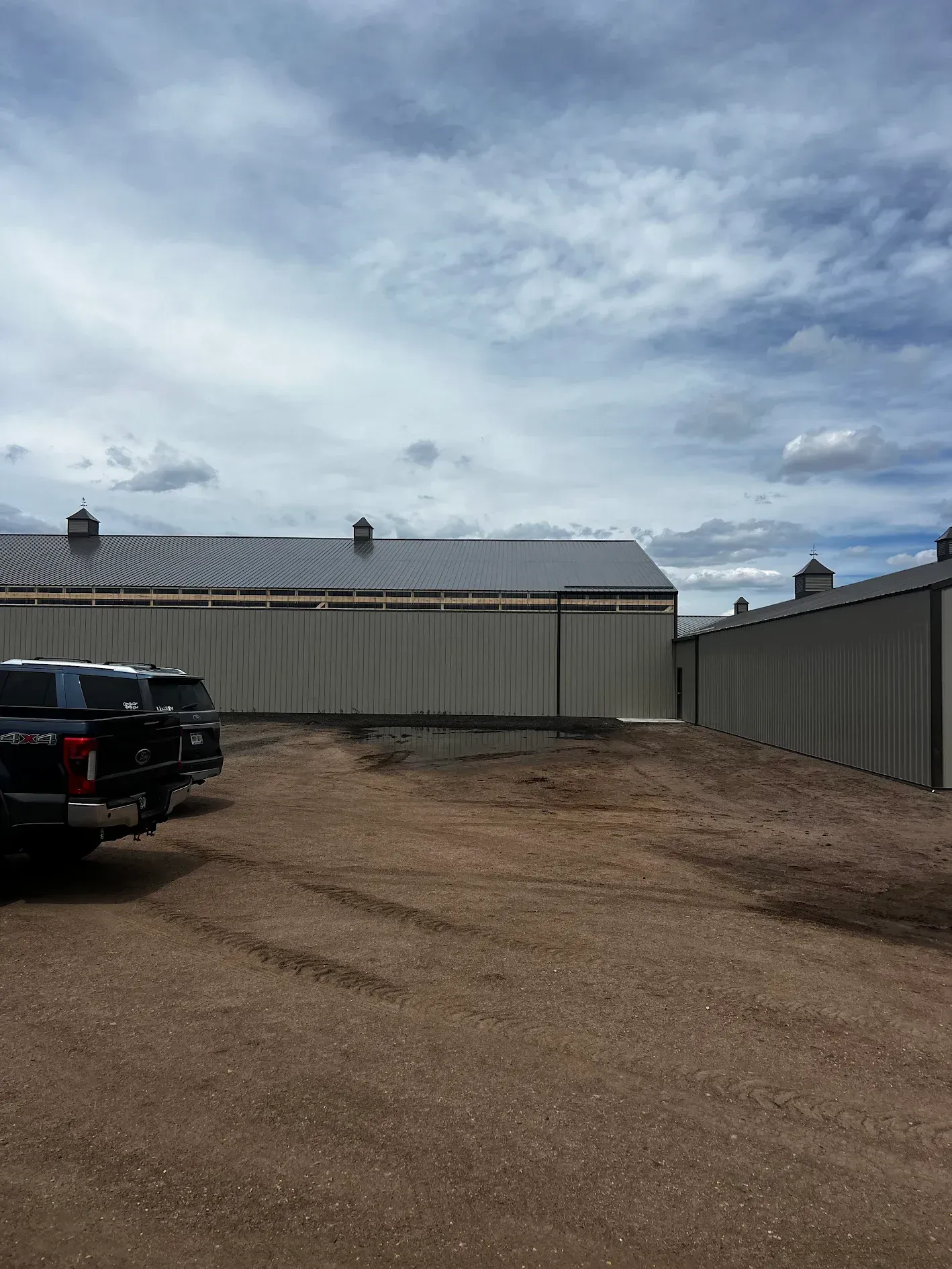 Gravel lot with a new building under construction; gray siding, black truck parked, cloudy sky.