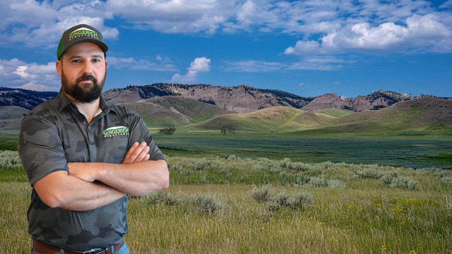 Bearded man in a cap stands with arms crossed in a grassy field with mountains and blue sky behind him
