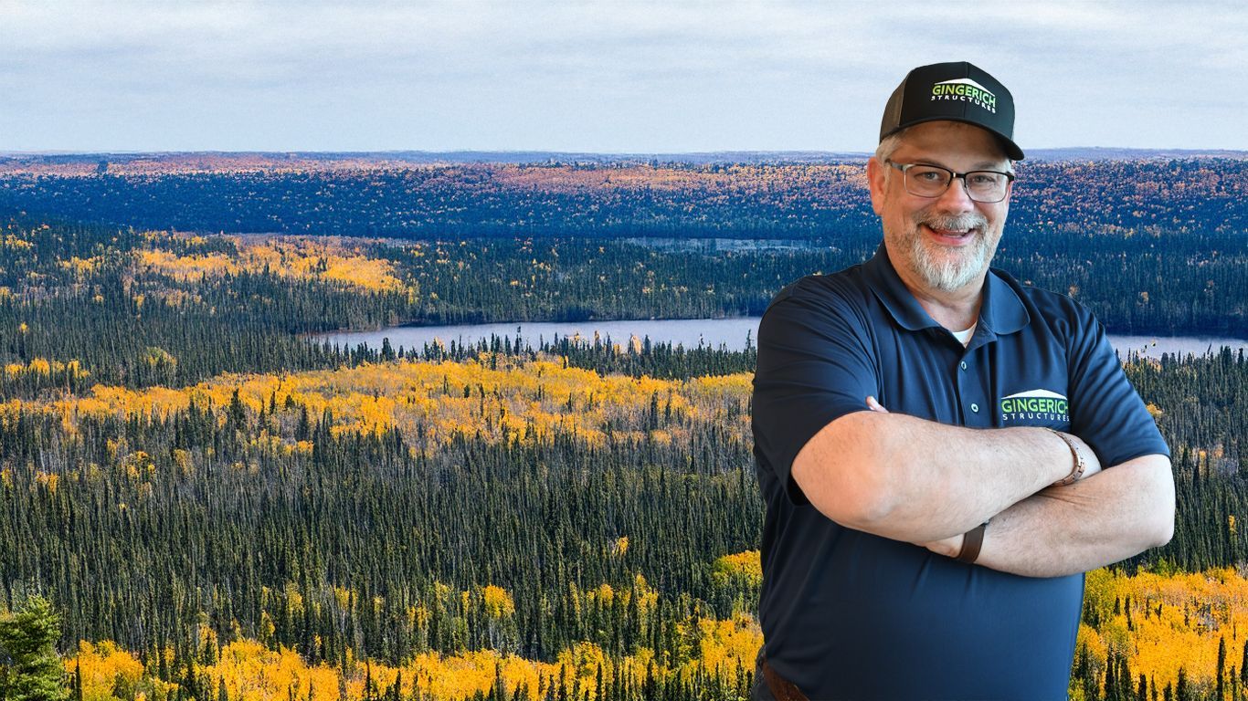 Smiling person with glasses and a cap, arms crossed, posing in front of a scenic autumn forest landscape with a lake.