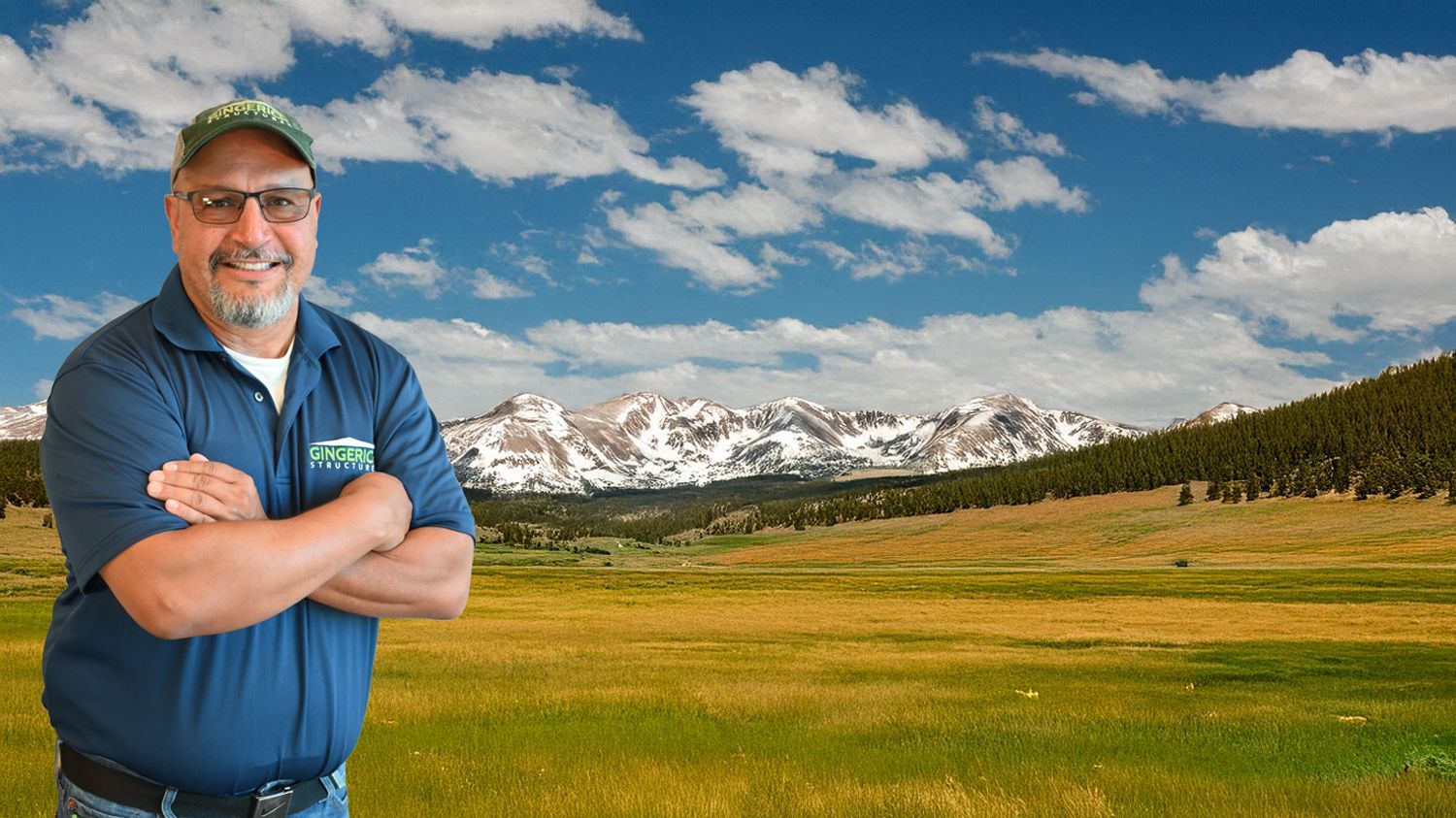 A smiling person in a blue polo shirt and cap stands with arms crossed against a backdrop of mountains and a field.