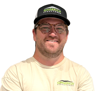Smiling man in a black cap and glasses, wearing a beige T-shirt, standing with arms crossed.