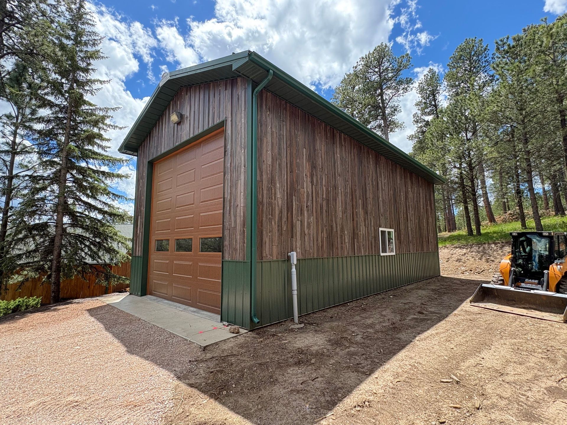 Large wooden barn with green trim and a closed garage door, beside a gravel yard and trees.