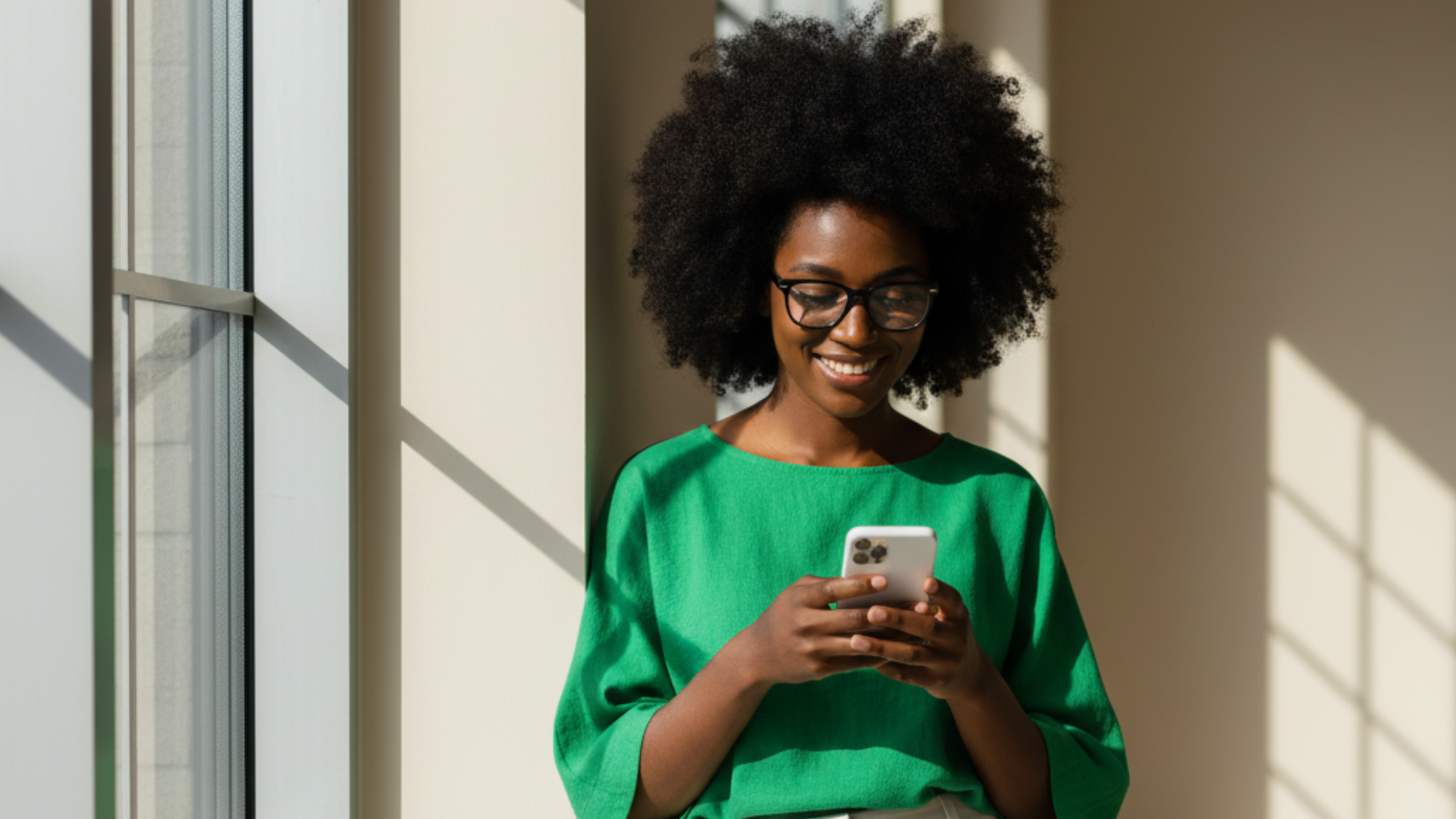 Une femme en chemise verte, portant des lunettes et une coiffure afro, sourit en regardant son téléphone devant une fenêtre.