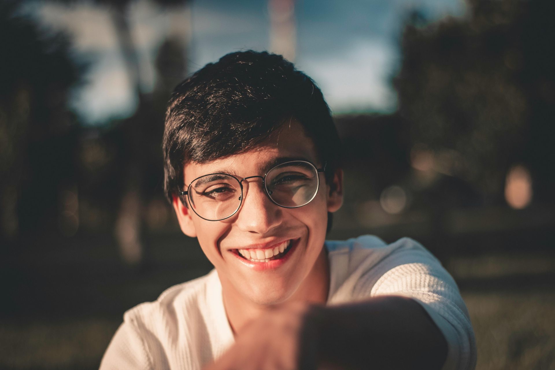 Personne souriante portant des lunettes et un haut blanc, en extérieur avec un arrière-plan flou.