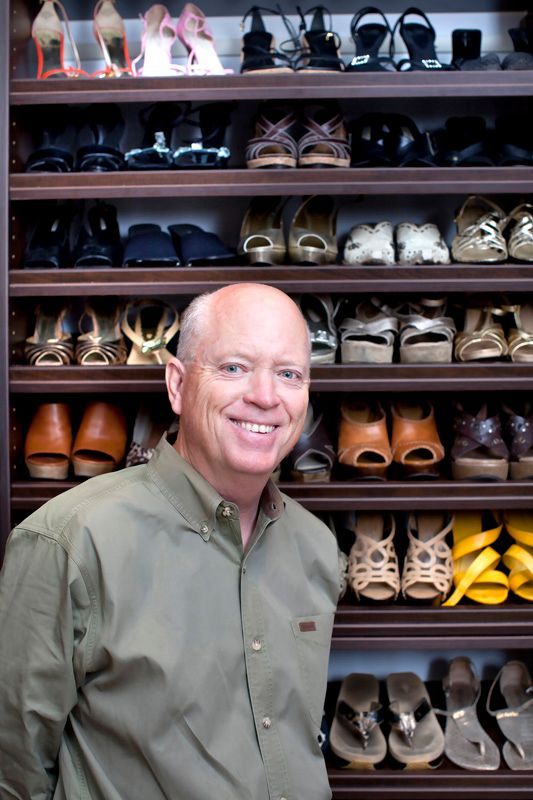 Man smiling, standing in front of shoe shelves filled with various women's shoes.