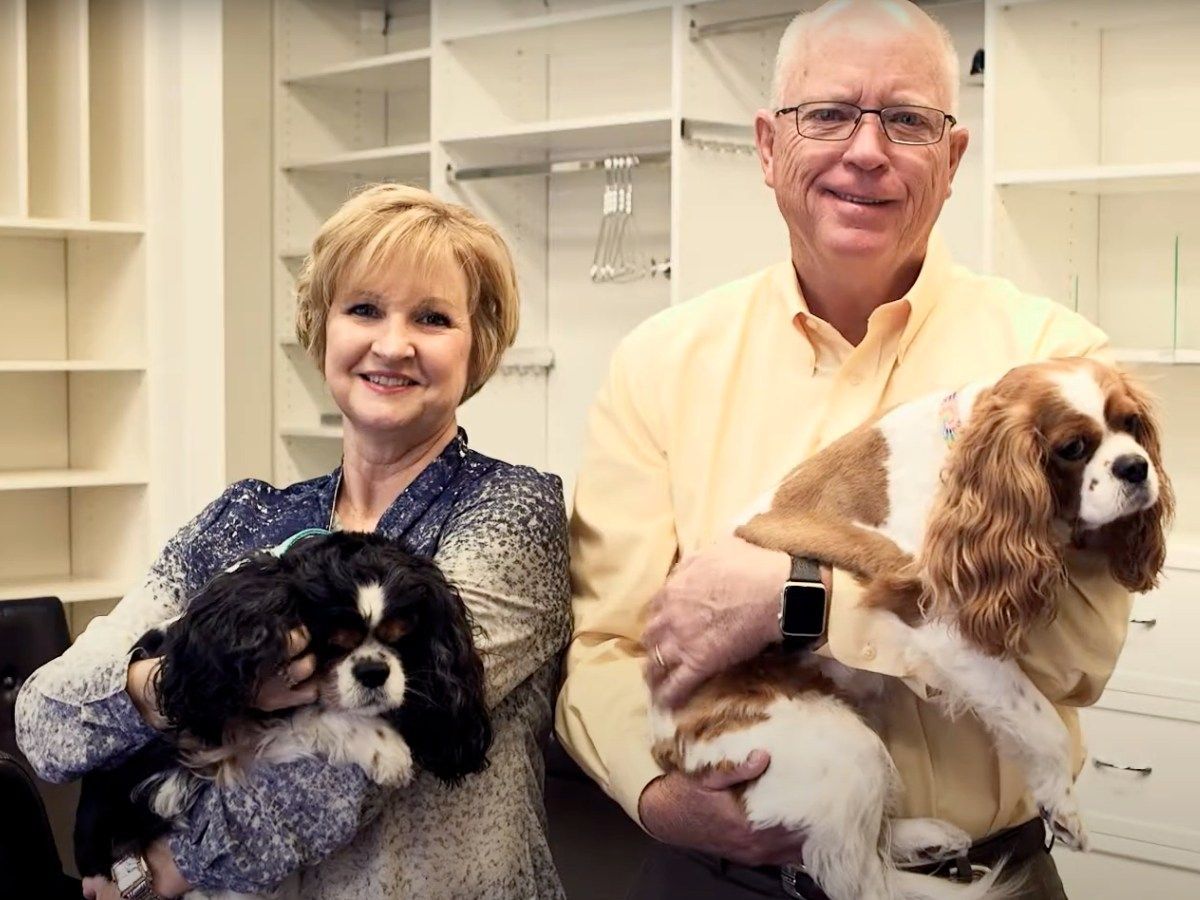 Woman and man smiling, each holding a Cavalier King Charles Spaniel dog in a closet.