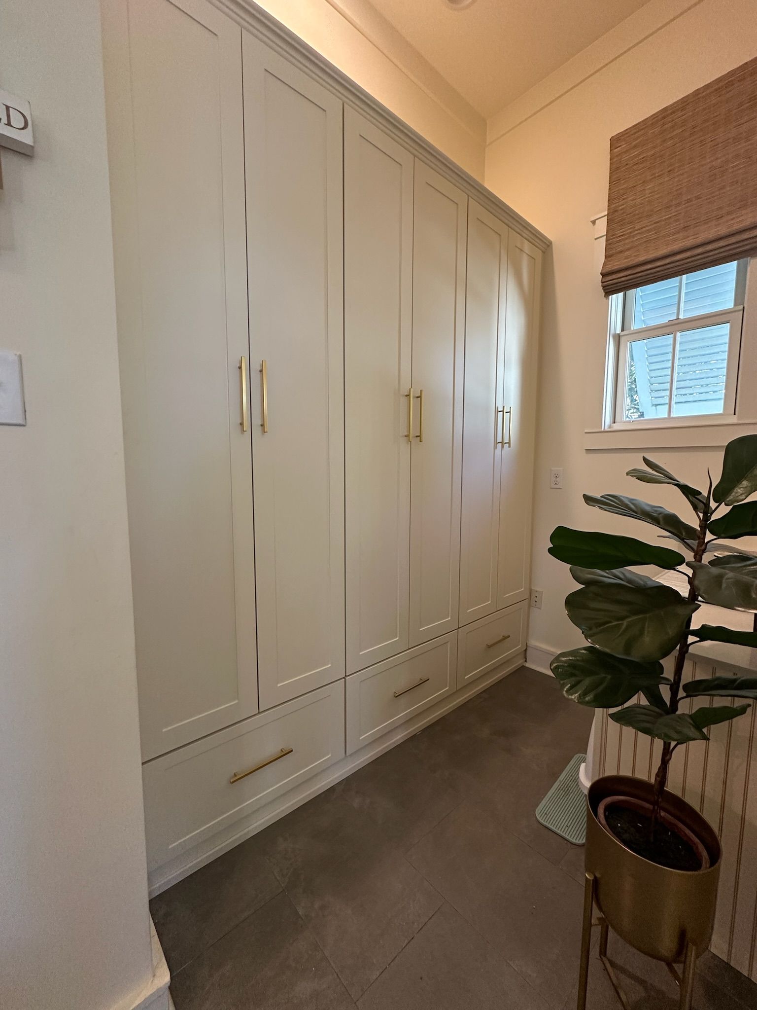 Built-in white storage cabinets with gold handles, next to a window with a woven shade and a potted plant.