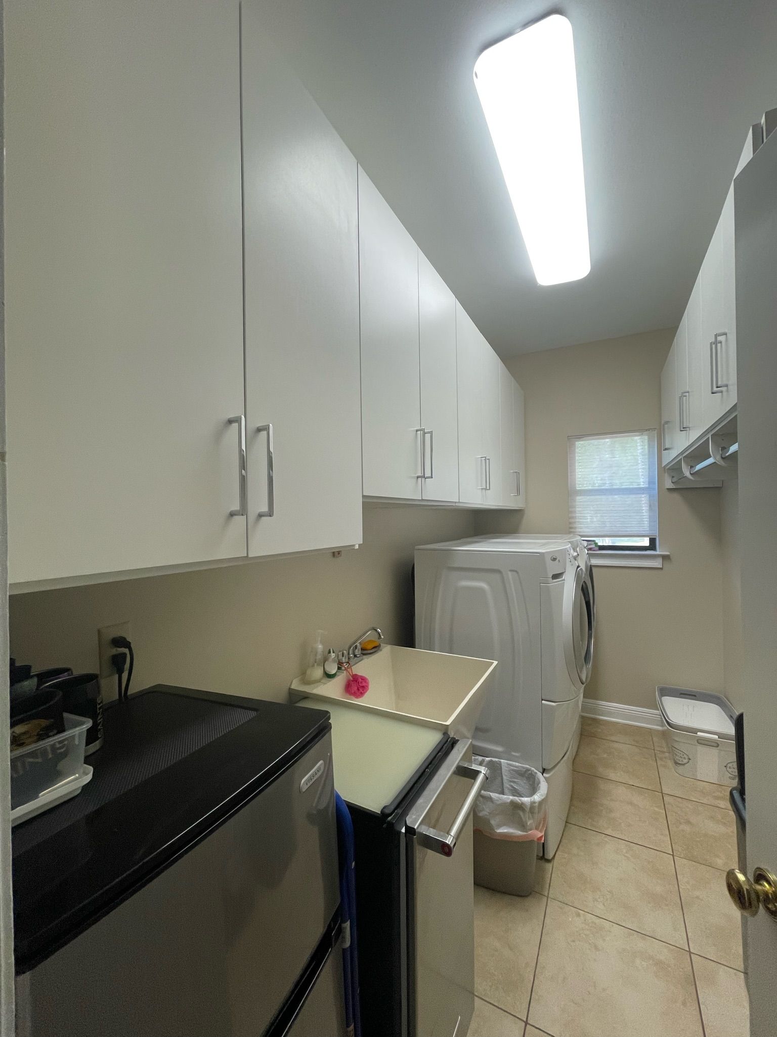 A white, narrow laundry room with overhead cabinets, a washer and dryer set, a utility sink, and tiled flooring.