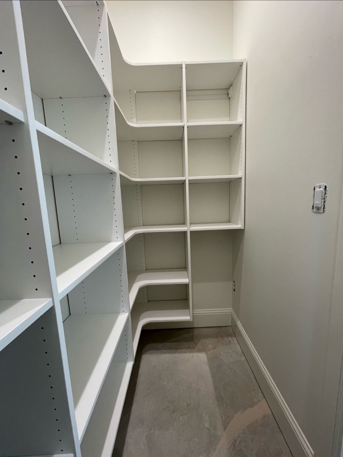 White built-in pantry shelving in a corner. Shelves of various sizes. Beige walls and cement floor.