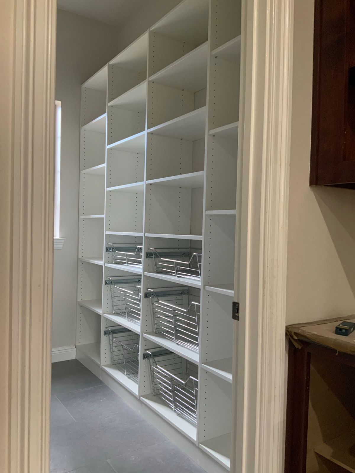White pantry shelving with pull-out wire baskets inside a doorway; gray flooring and a window are visible.