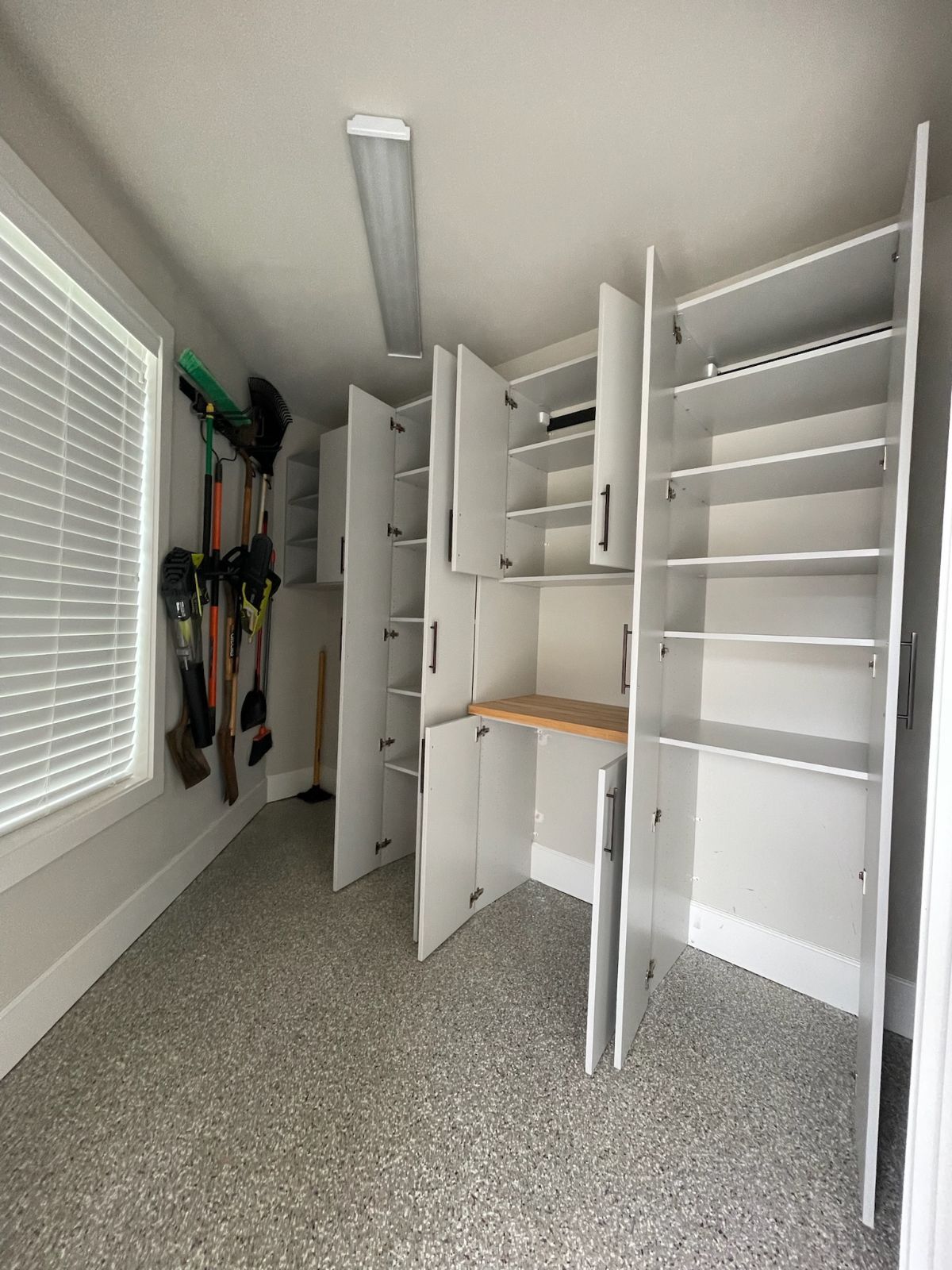 Garage interior with white cabinets and shelving, tools hanging, and speckled gray flooring.