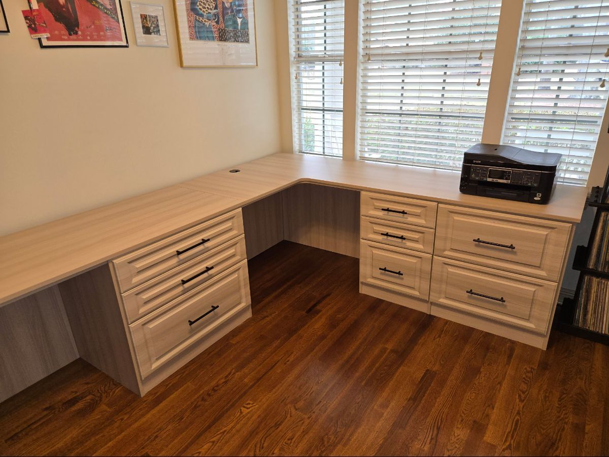 L-shaped desk with drawers by a window. Light wood desk, dark wood floor, printer.