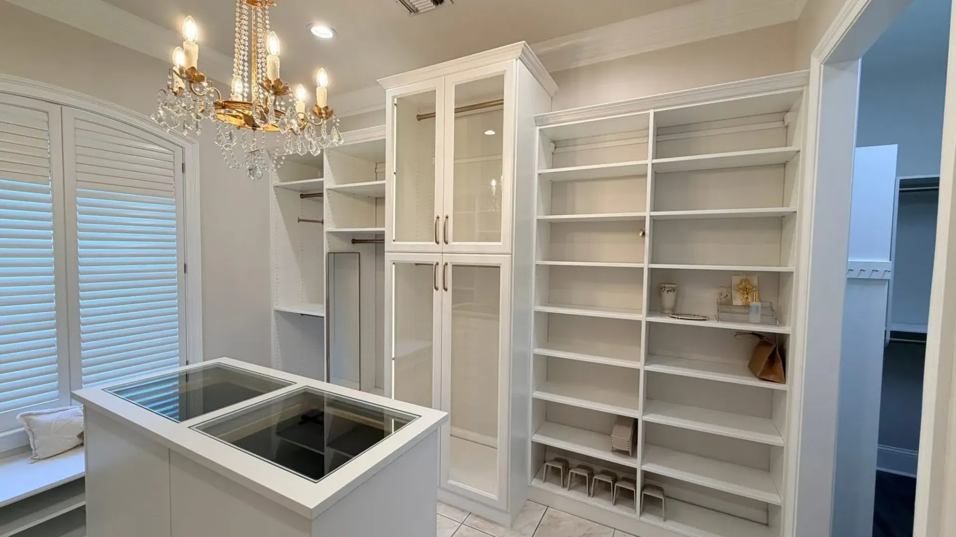 A white walk-in closet featuring floor-to-ceiling shelving, a glass-front cabinet, a central island, and a chandelier.