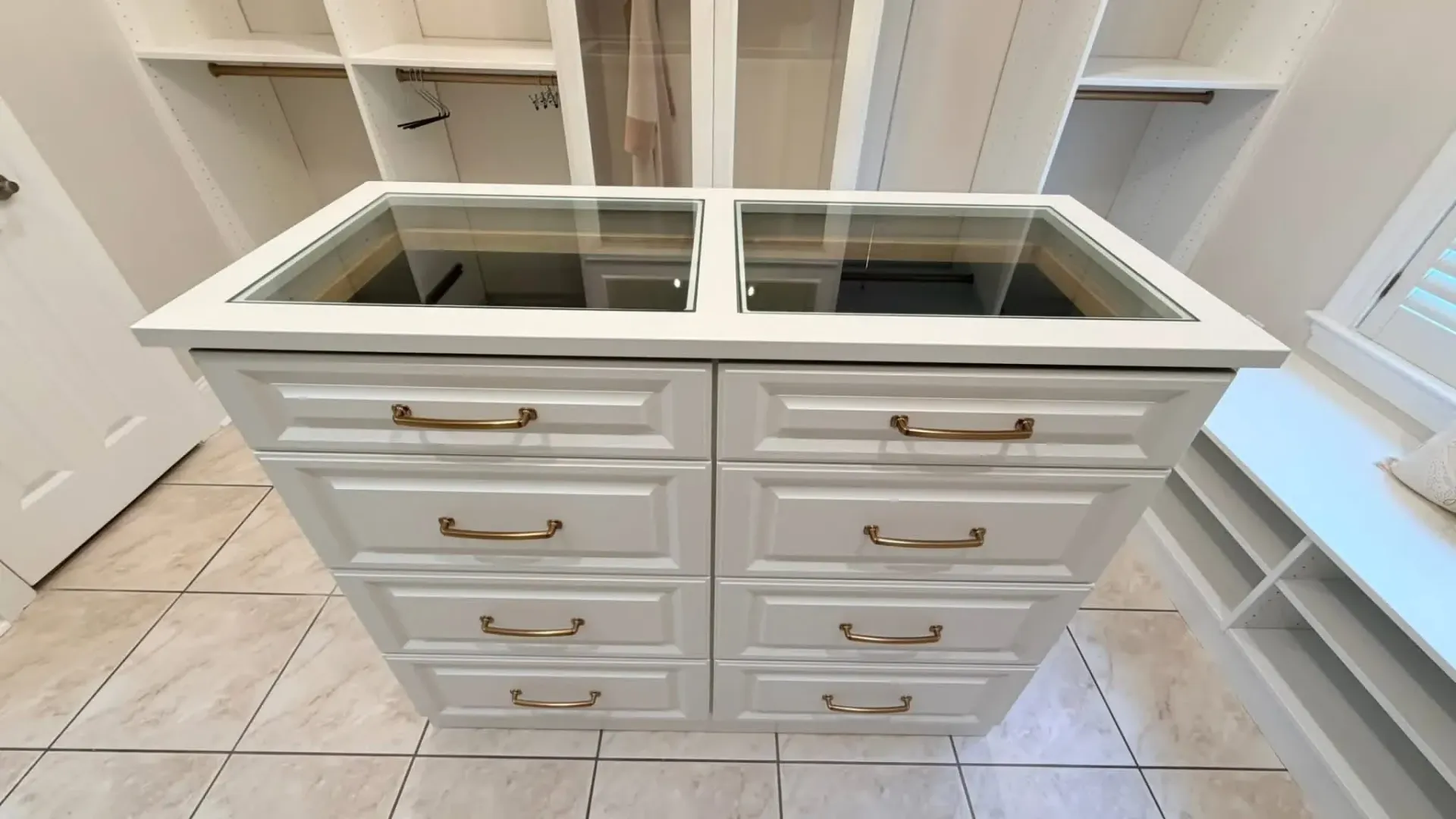 A white wooden closet island with eight drawers and a glass top sitting in the center of a walk-in closet.