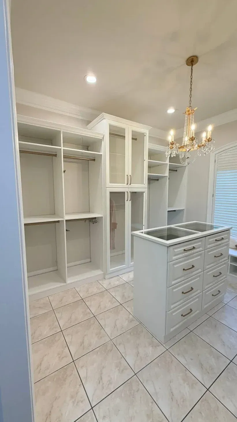 A white walk-in closet featuring floor-to-ceiling storage, a central island with drawers, and a gold crystal chandelier.