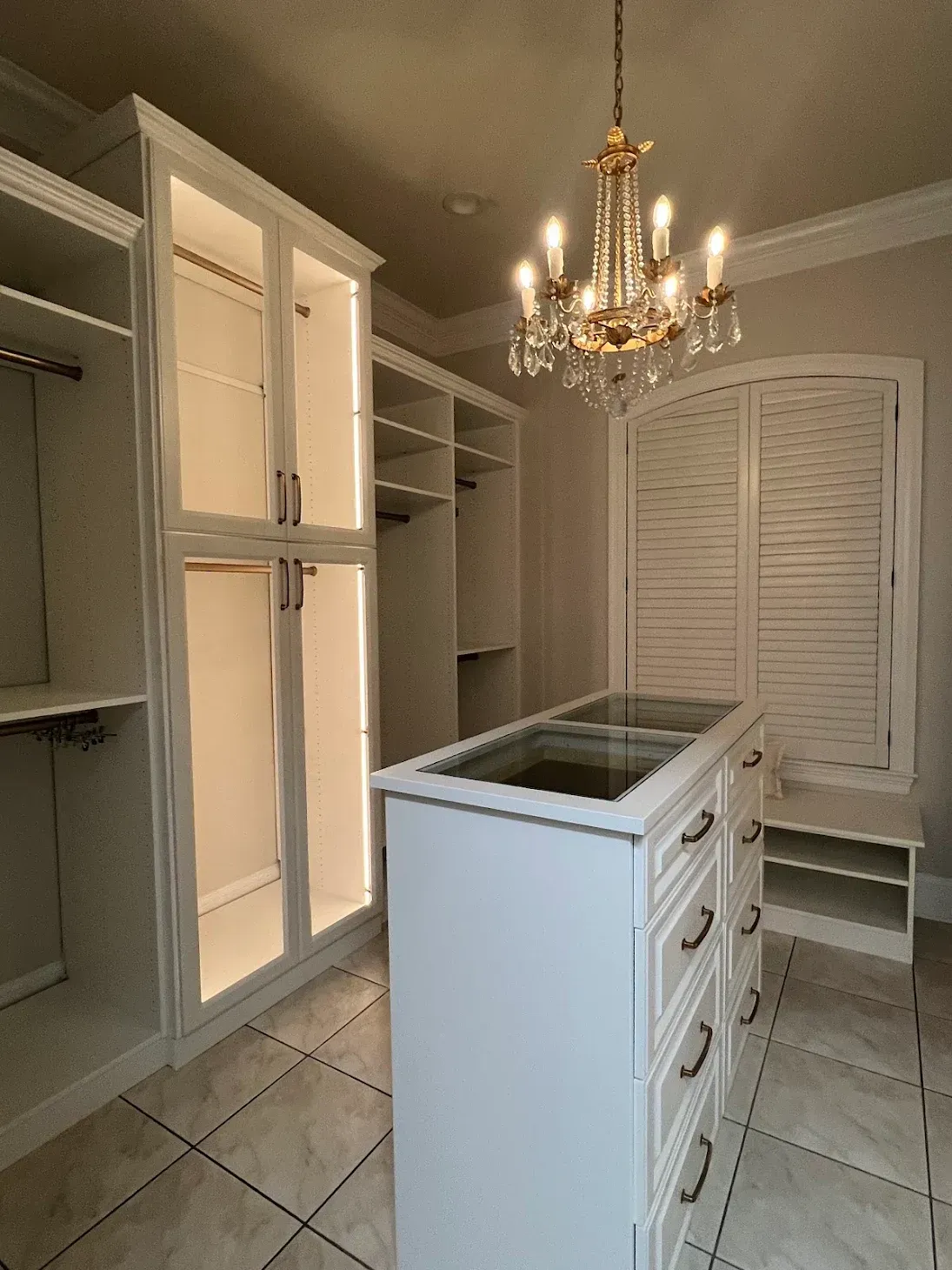 A walk-in closet featuring white cabinets with interior lighting, a glass-topped storage island, and a crystal chandelier.