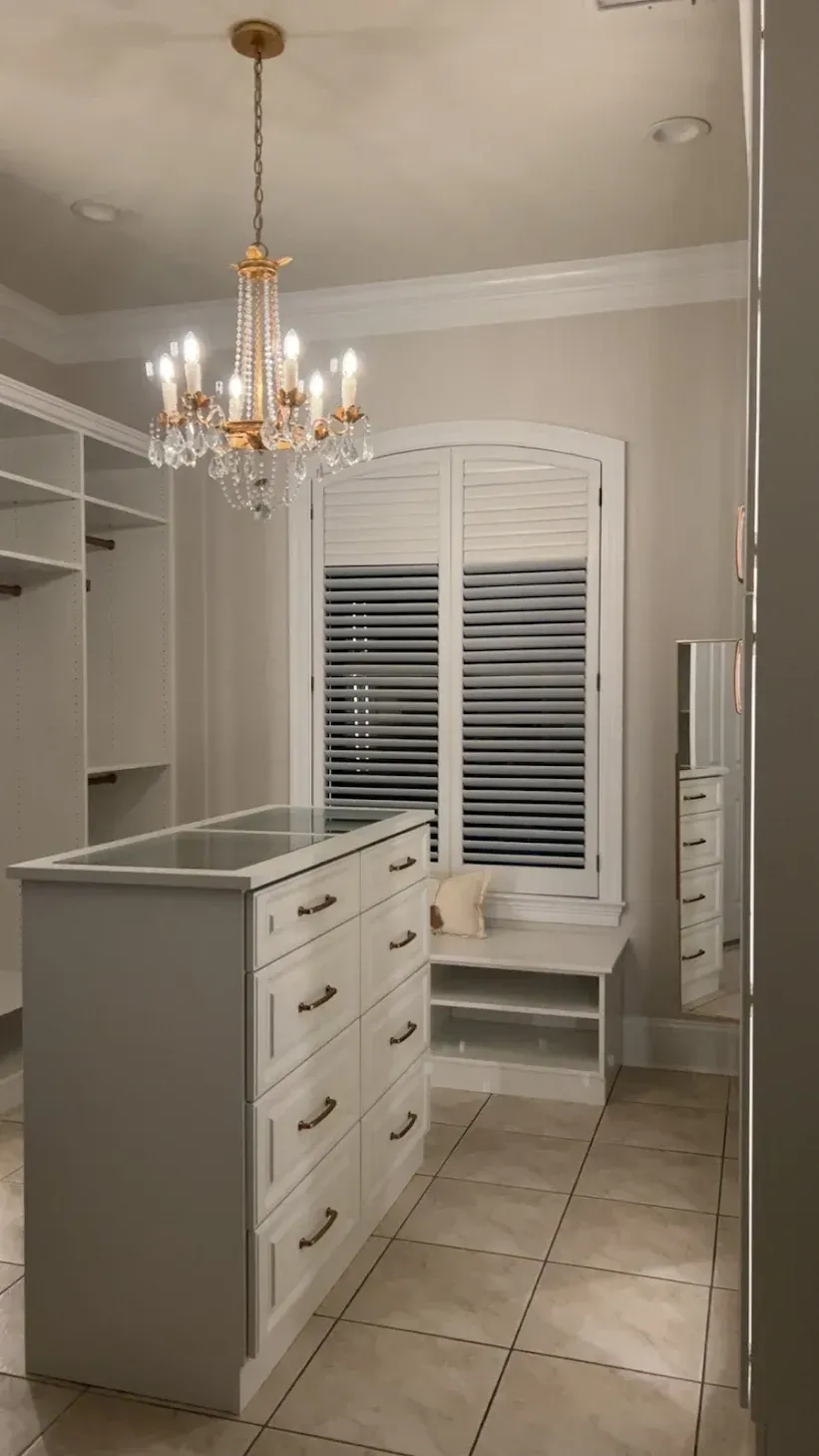 A bright, organized walk-in closet featuring a central white dresser, shelving units, a chandelier, and a window seat.