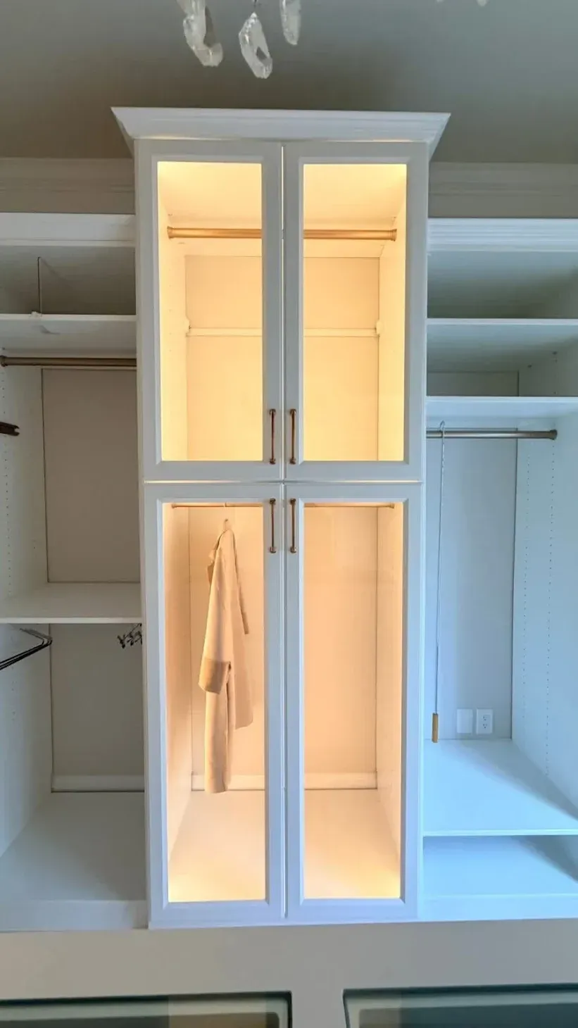 White custom closet unit with central glass-front cabinets featuring interior LED lighting and open side shelving.