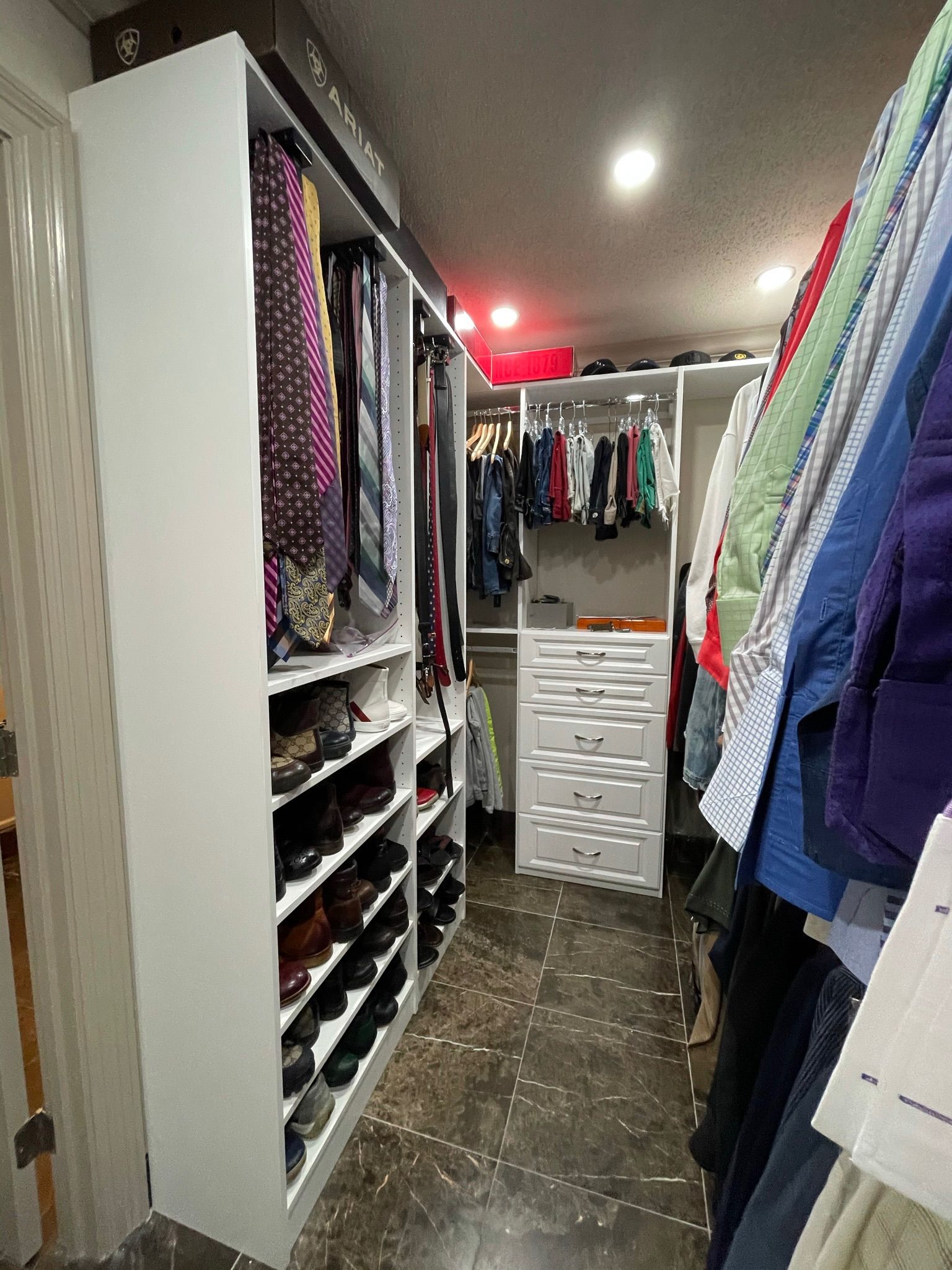 Walk-in closet with white shelves and drawers, filled with hanging clothes, shoes, and accessories.