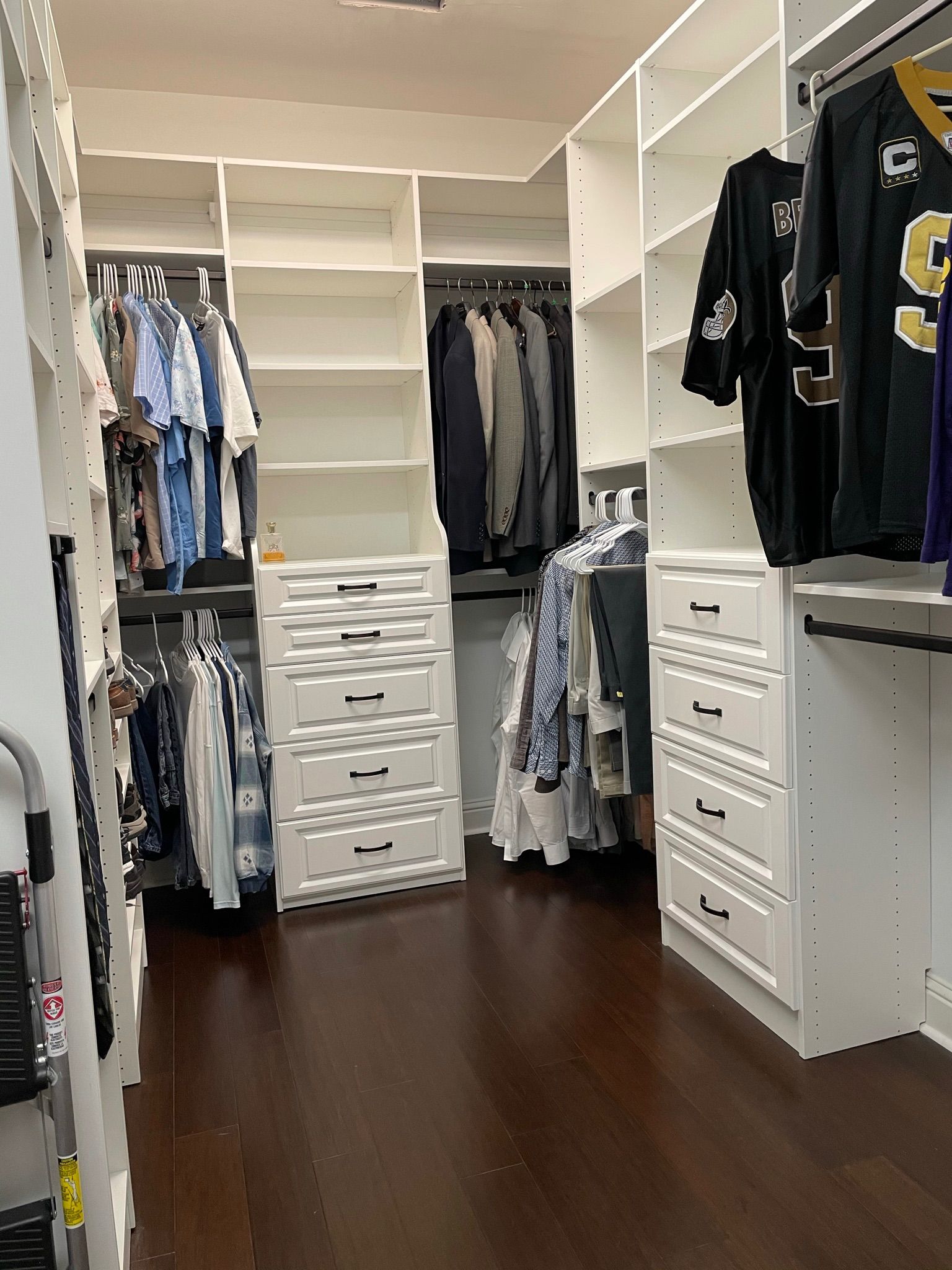 Walk-in closet with white shelves and drawers, dark wood floor, clothes hanging, and a New Orleans Saints jersey.