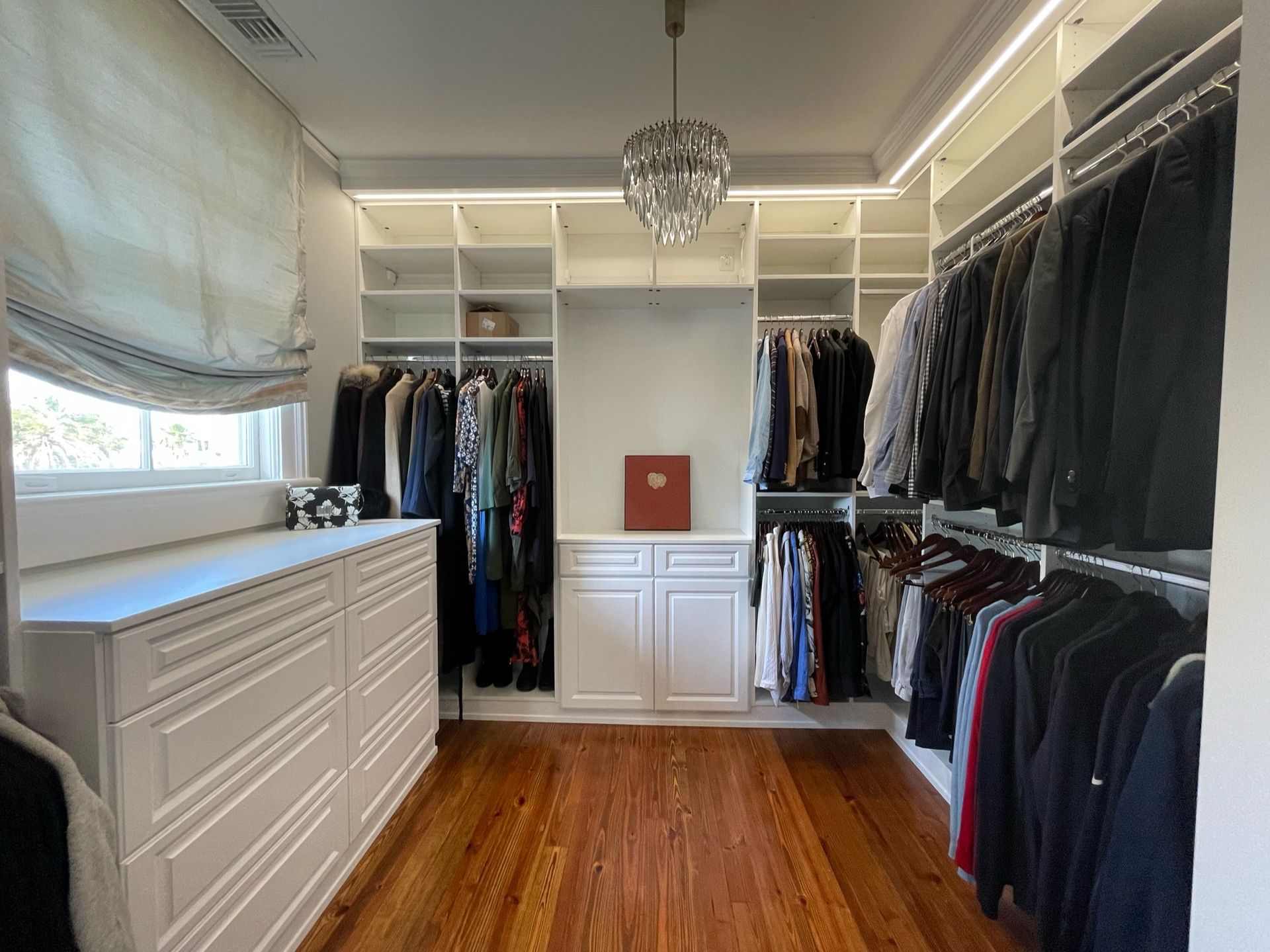 Walk-in closet with white shelves and drawers, filled with hanging clothes. Wooden floor and chandelier.