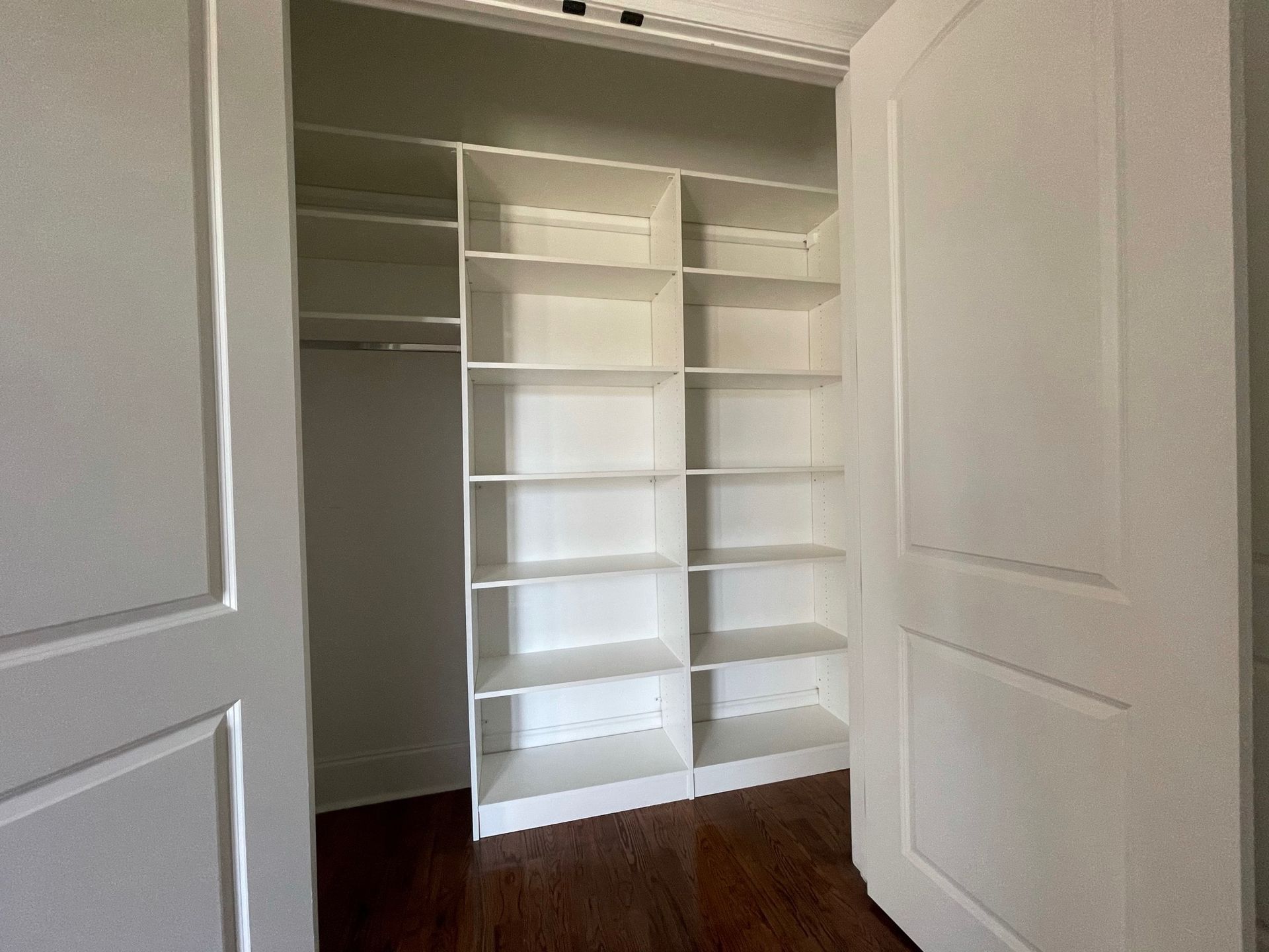 Empty white closet with built-in shelving and a hanging rod. Wood floor, white doors, and shelves.