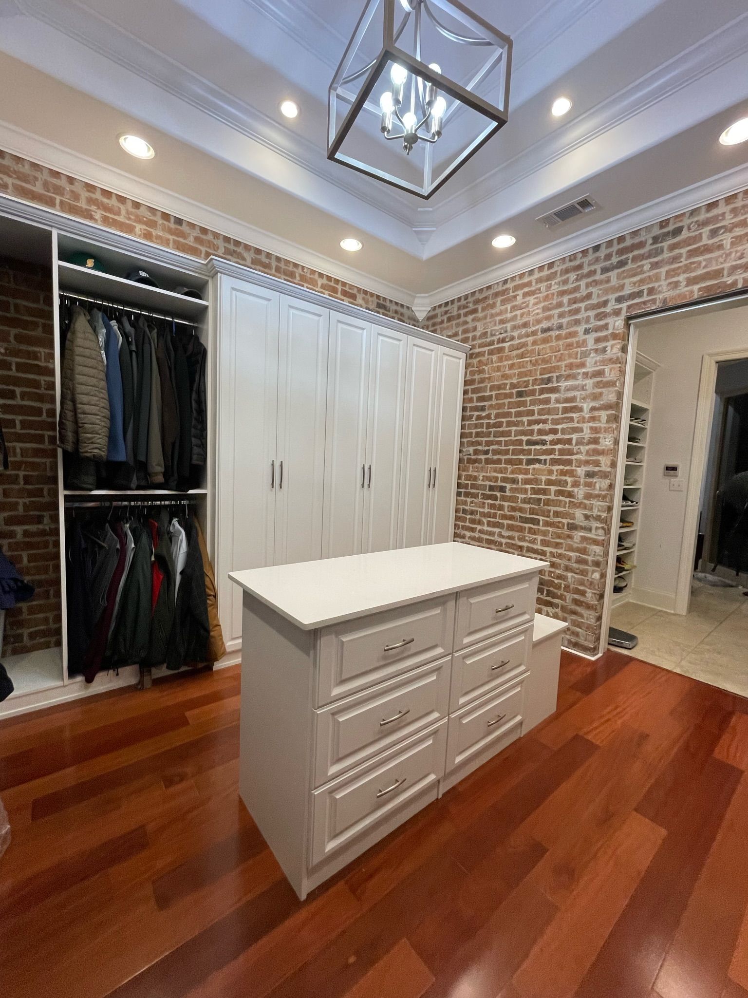 Walk-in closet with red brick wall, white cabinets, and wooden floor. Clothes hang on the left.