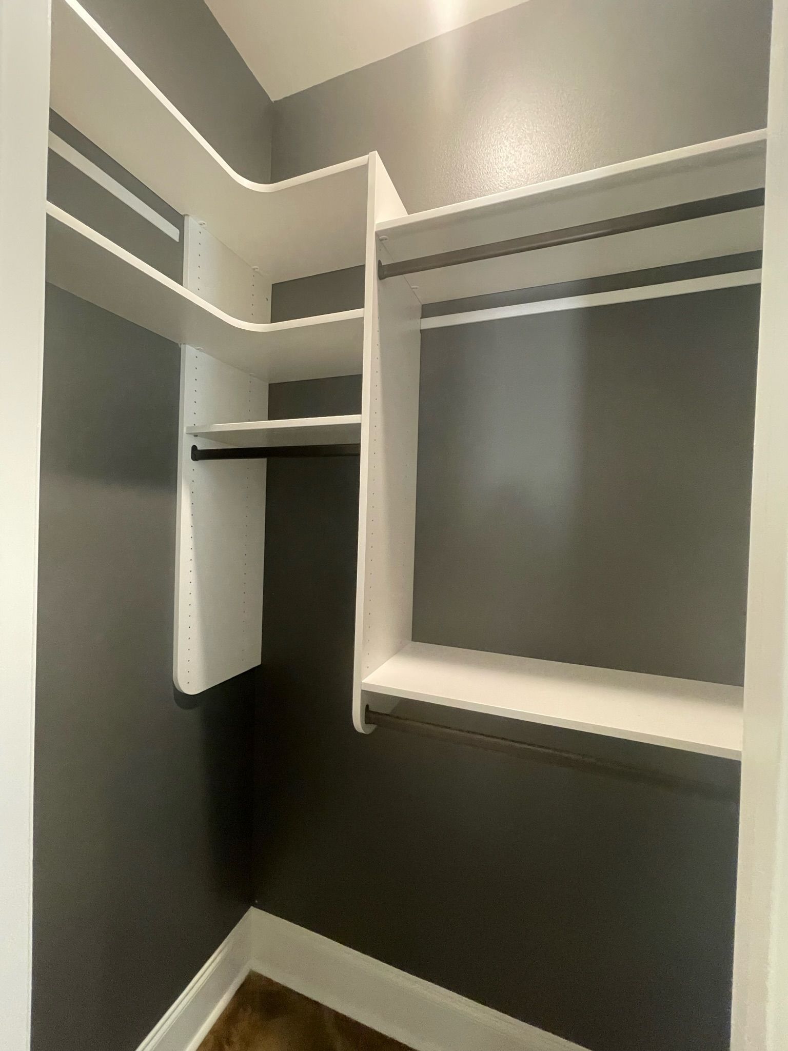 Empty closet with white shelving and hanging rods; gray walls and brown floor.