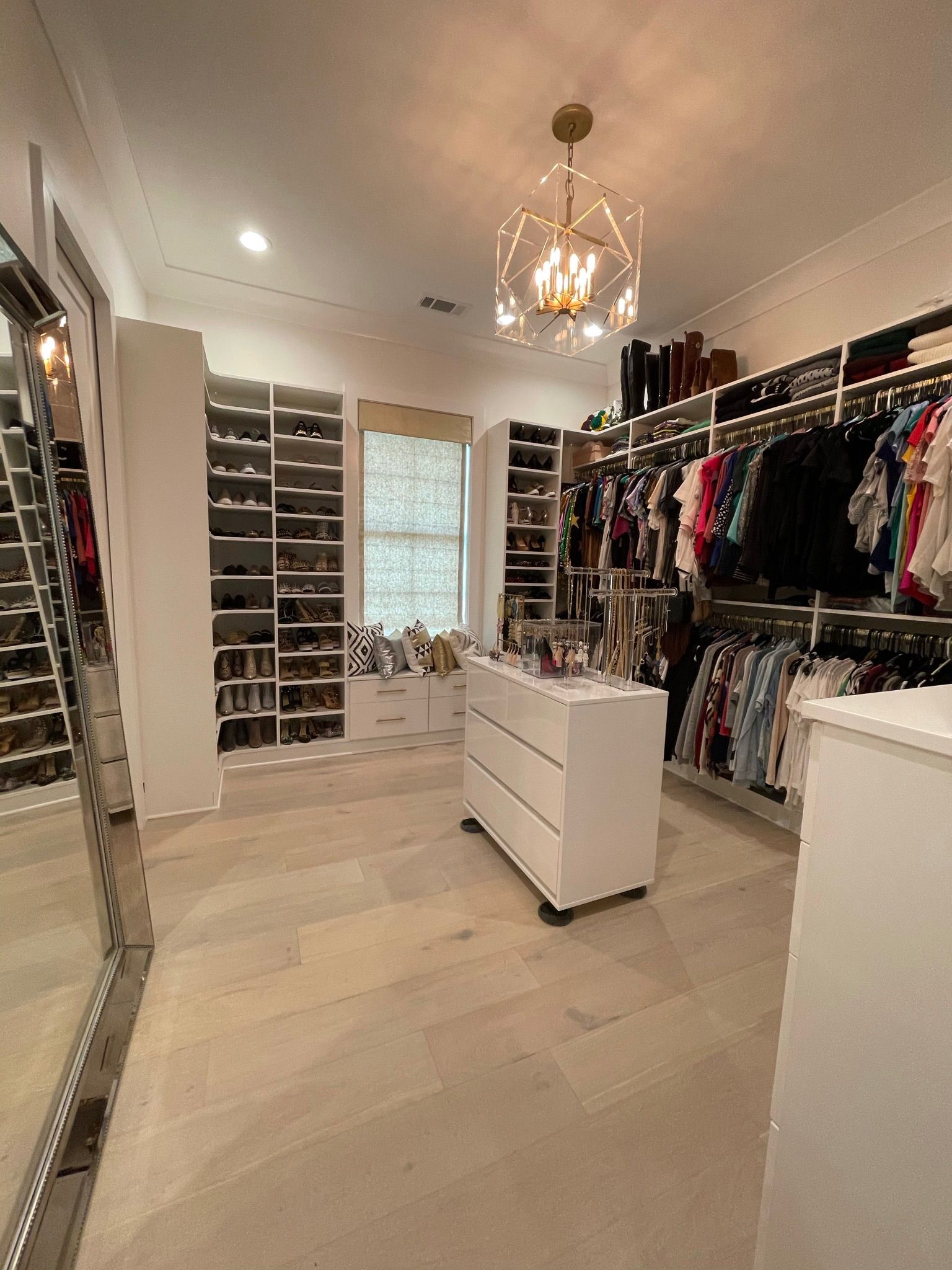 Walk-in closet with white shelves, clothing racks, and a central dresser. Light wood floors, bright lighting.