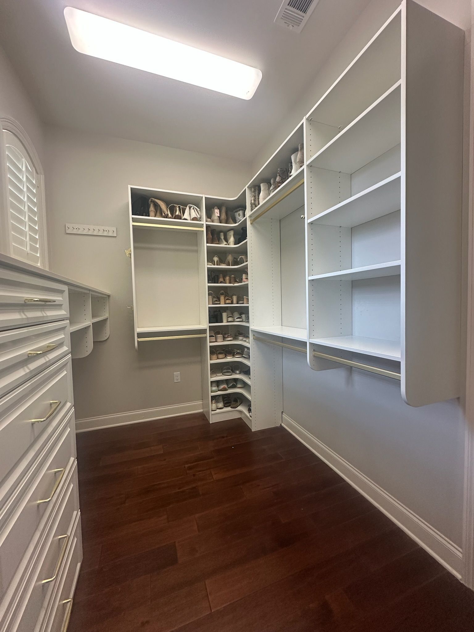 Walk-in closet with white shelves and drawers, dark wood floor, and overhead lighting.