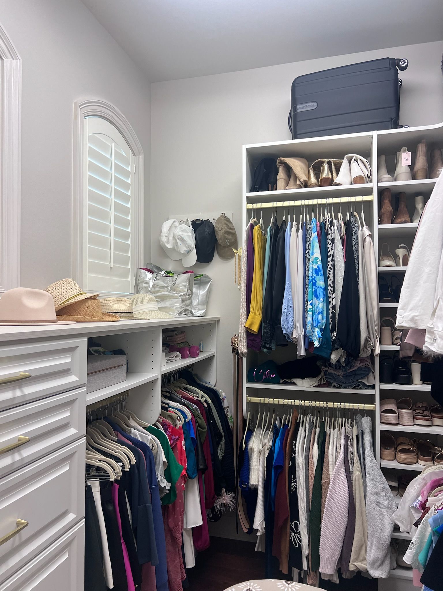 Walk-in closet with hanging clothes, shelves for shoes and accessories, and a small window with shutters.