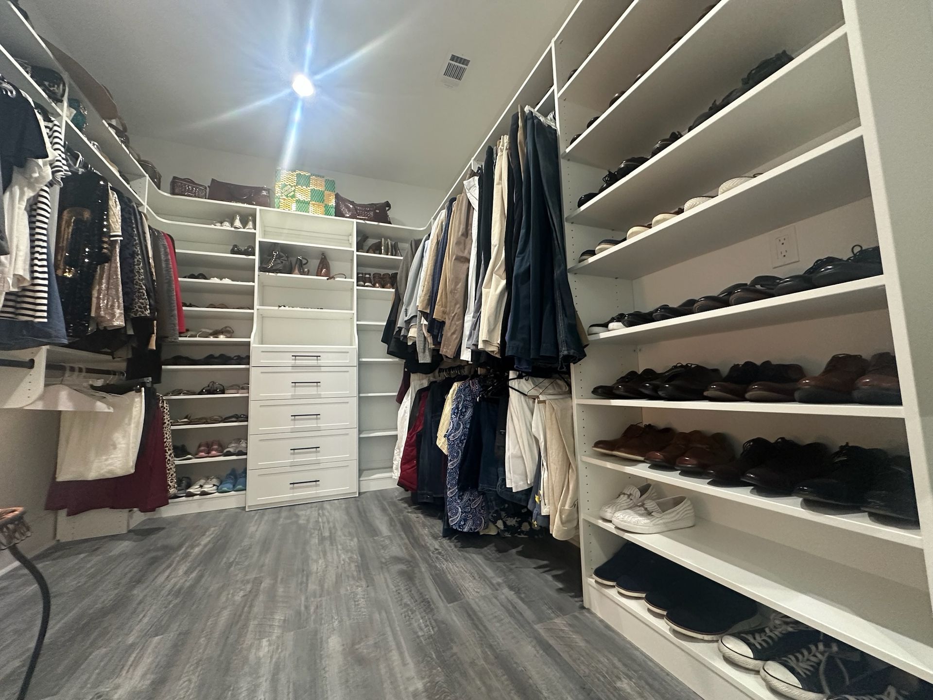 Walk-in closet with white shelving, displaying shoes and hanging clothes. Gray flooring.