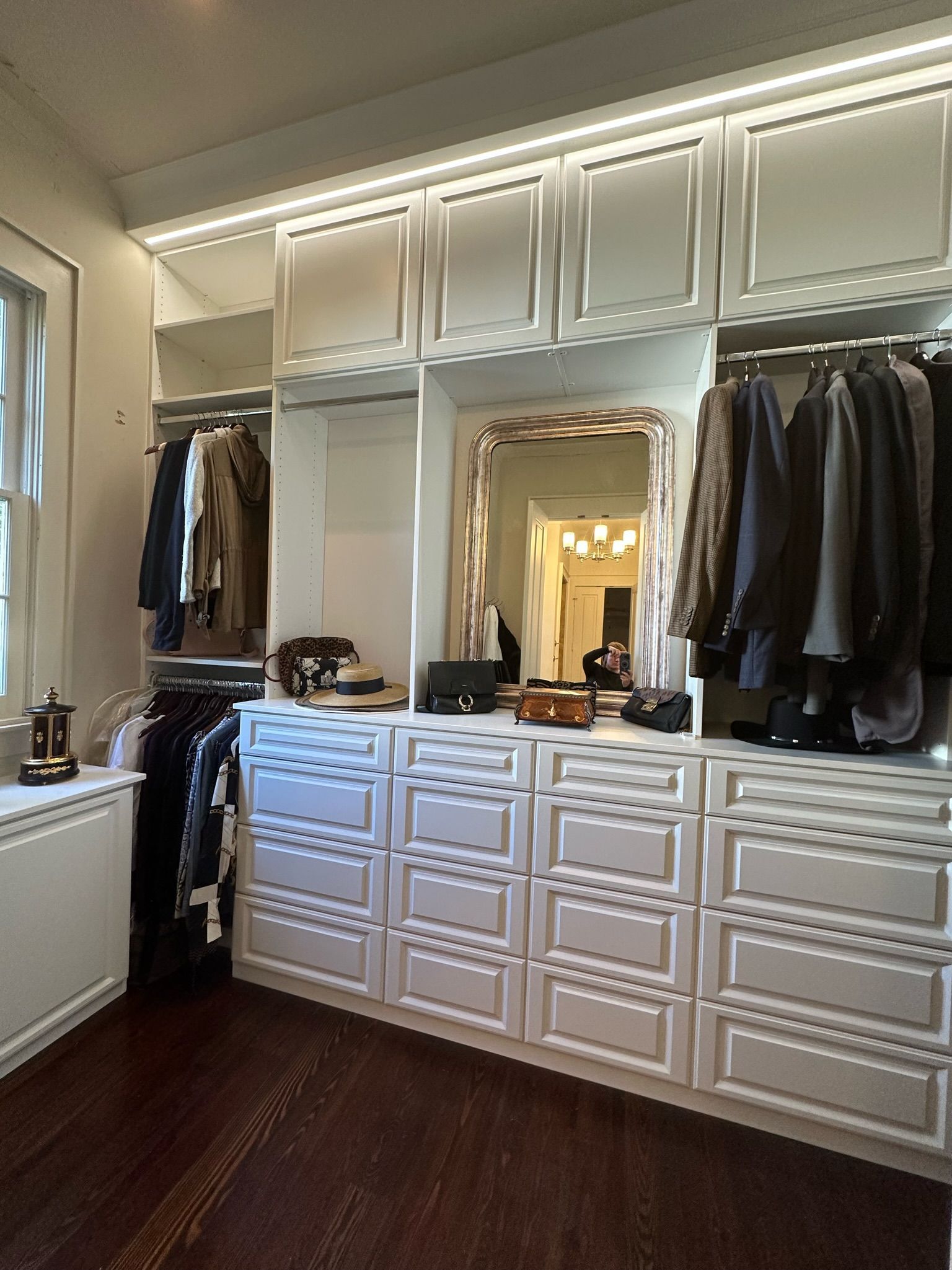 White walk-in closet with drawers, hanging clothes, and a large mirror; hardwood floor, built-in lights.