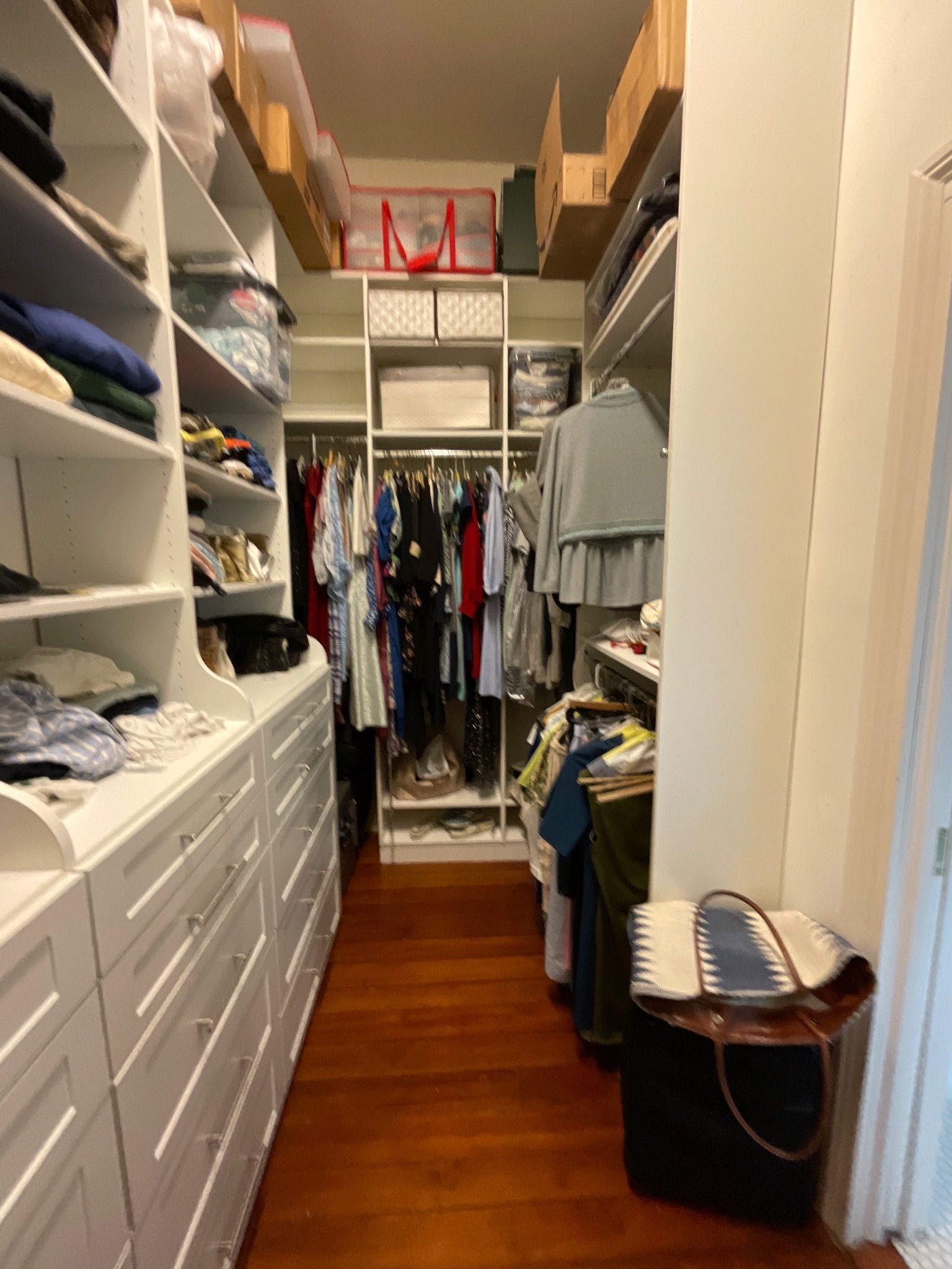 Narrow walk-in closet with white shelves and drawers, filled with clothes. Dark wood floor.