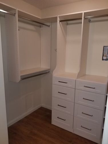 White closet with drawers, shelves, and hanging rods in a corner room, on wooden floor.