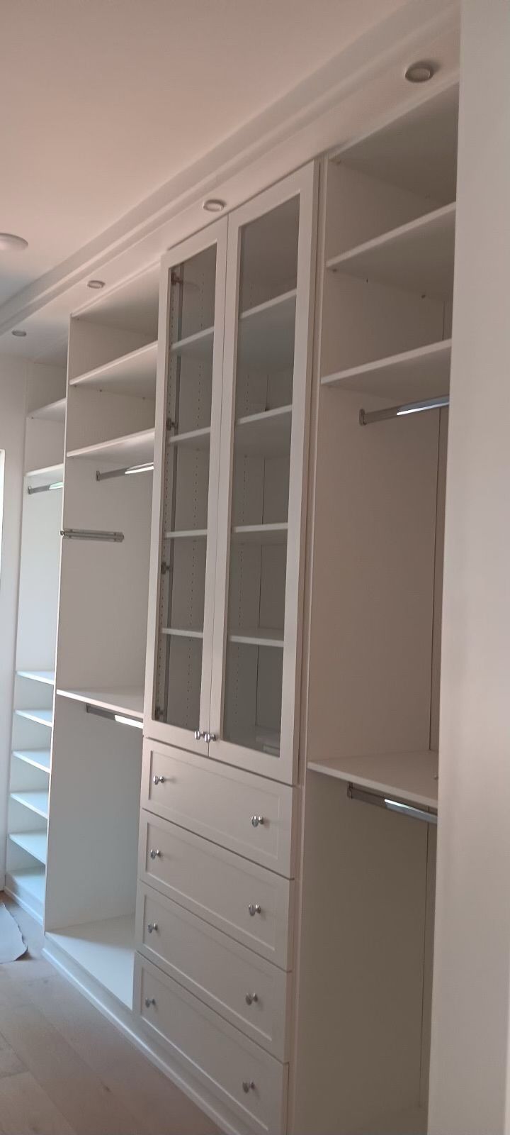 White built-in closet with shelves, drawers, and glass-door cabinets. Recessed lighting in the ceiling.
